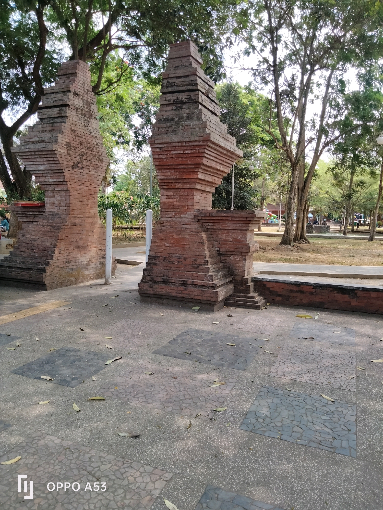 The image captures an outdoor scene in the city of Trenggalek, Indonesia, focusing on a pair of ornate brick gateways. These structures, built from reddish-brown bricks, feature stepped designs and are integrated into a park-like environment. The foreground consists of a paved area with patterned concrete tiles and scattered fallen leaves. Beyond the gateways, a pathway leads into a grassy area populated by several large trees with sparse foliage, suggesting a dry season. In the background, more trees and foliage are visible, along with glimpses of park amenities and a few people engaged in activities such as sitting and walking. The lighting indicates it is daytime, likely late morning or early afternoon, with the sun casting shadows through the trees. The overall impression is of a serene public space within Trenggalek.