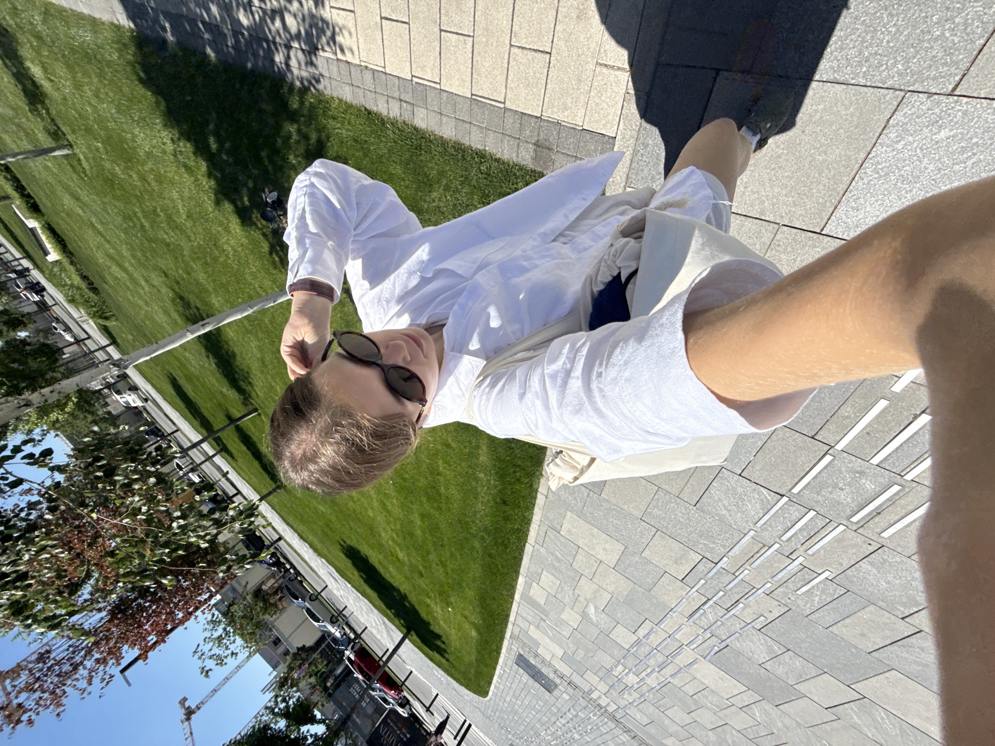 A selfie taken at ground level depicts a person wearing sunglasses and a white button-down shirt. The photo is angled, with the person appearing to stand on a light gray brick walkway bordered by green grass. Buildings, including a crane, are visible in the background, suggesting an urban area in Belgrade, Serbia.