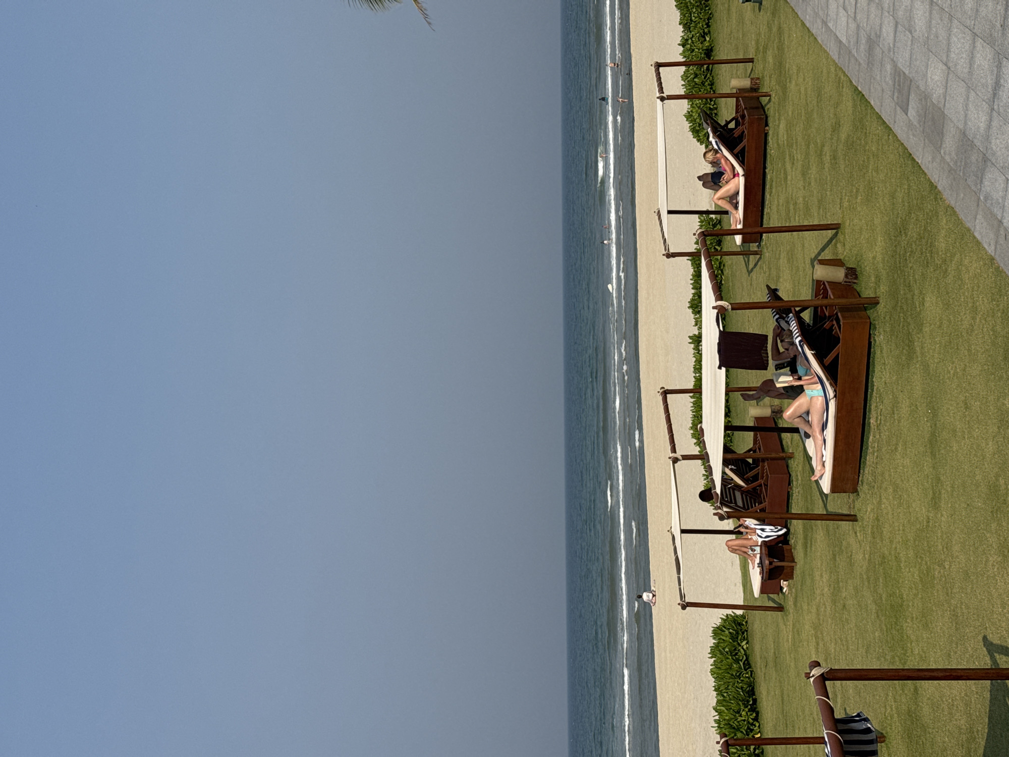 The image depicts a beach scene in Đà Nẵng, Vietnam. The sky is clear and blue, transitioning to the ocean, which is a lighter blue. The shoreline is a sandy beach with gentle waves lapping the shore. A row of wooden cabanas with canvas shades are situated on a green lawn adjacent to the beach. People are lounging on the cabanas, some reading. Towels and personal belongings are visible within the cabanas. The lawn borders a paved area.