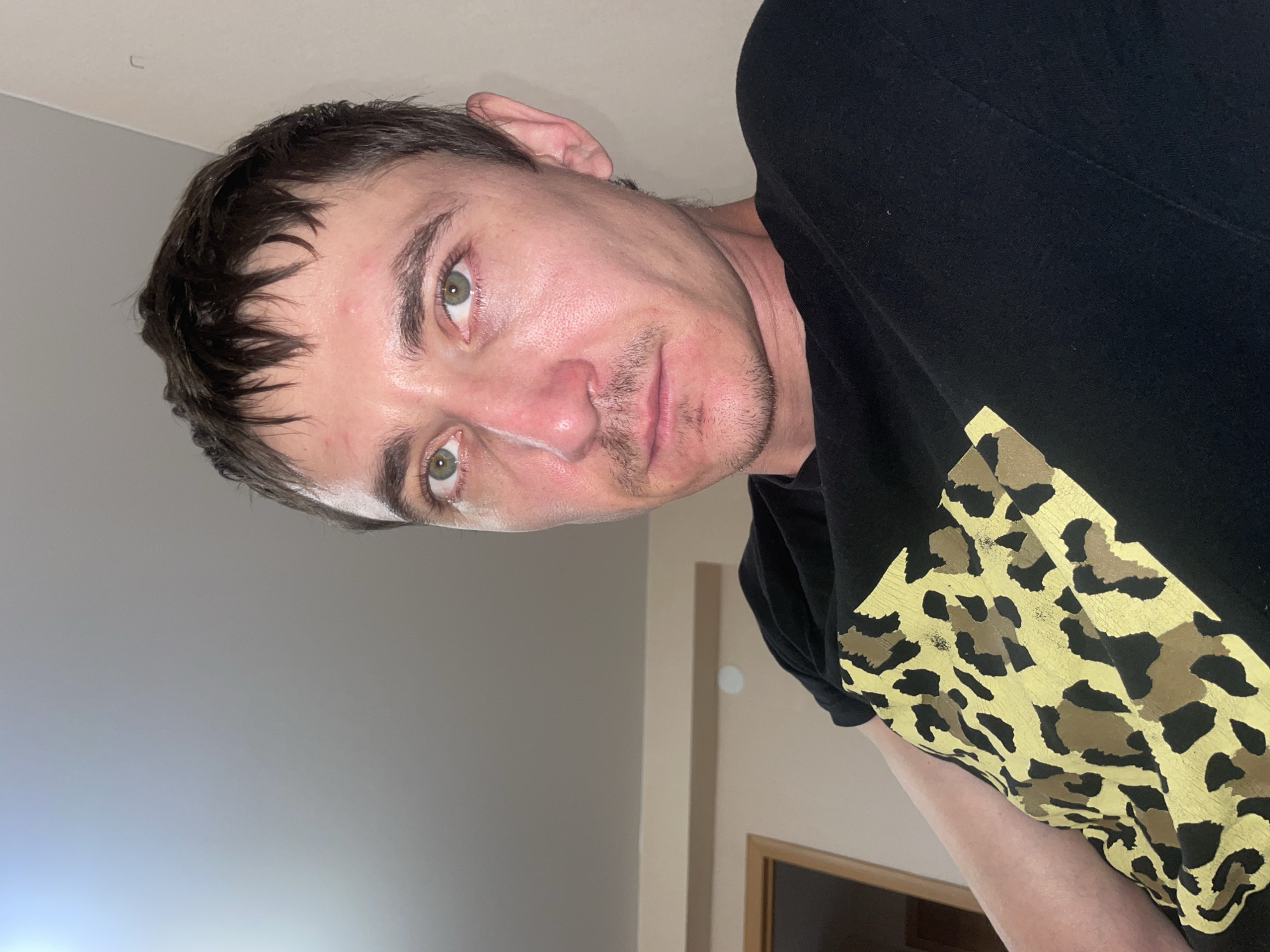 This is a medium close-up, head-and-shoulders selfie taken by a man indoors. The man has light skin, short dark brown hair, and green eyes. He is wearing a black t-shirt with a gold and black leopard print design. He has a light mustache and stubble. The background is out of focus and appears to be a room with a light-colored wall and a ceiling. The lighting is soft and appears to be coming from above and to the left. There is a doorway visible in the background on the left. The image was taken in Laishevo, Russia.