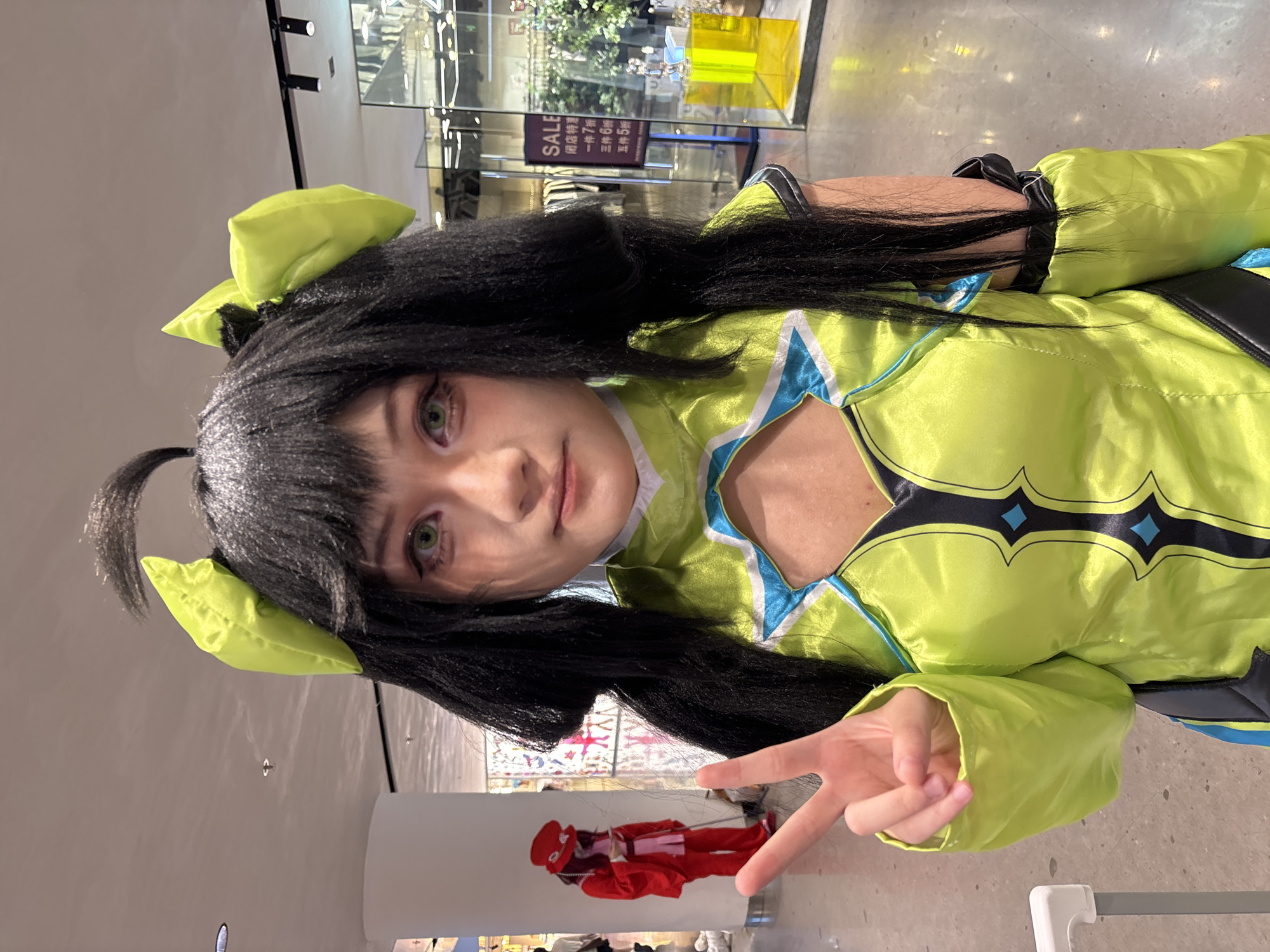 The image shows a woman in a green cosplay outfit with black hair styled with green accents, posing with a peace sign. She appears to be indoors in a commercial space, possibly a shopping mall or department store, as indicated by the presence of display windows and sale signage in the background. A mannequin dressed in red is visible through the window behind her.