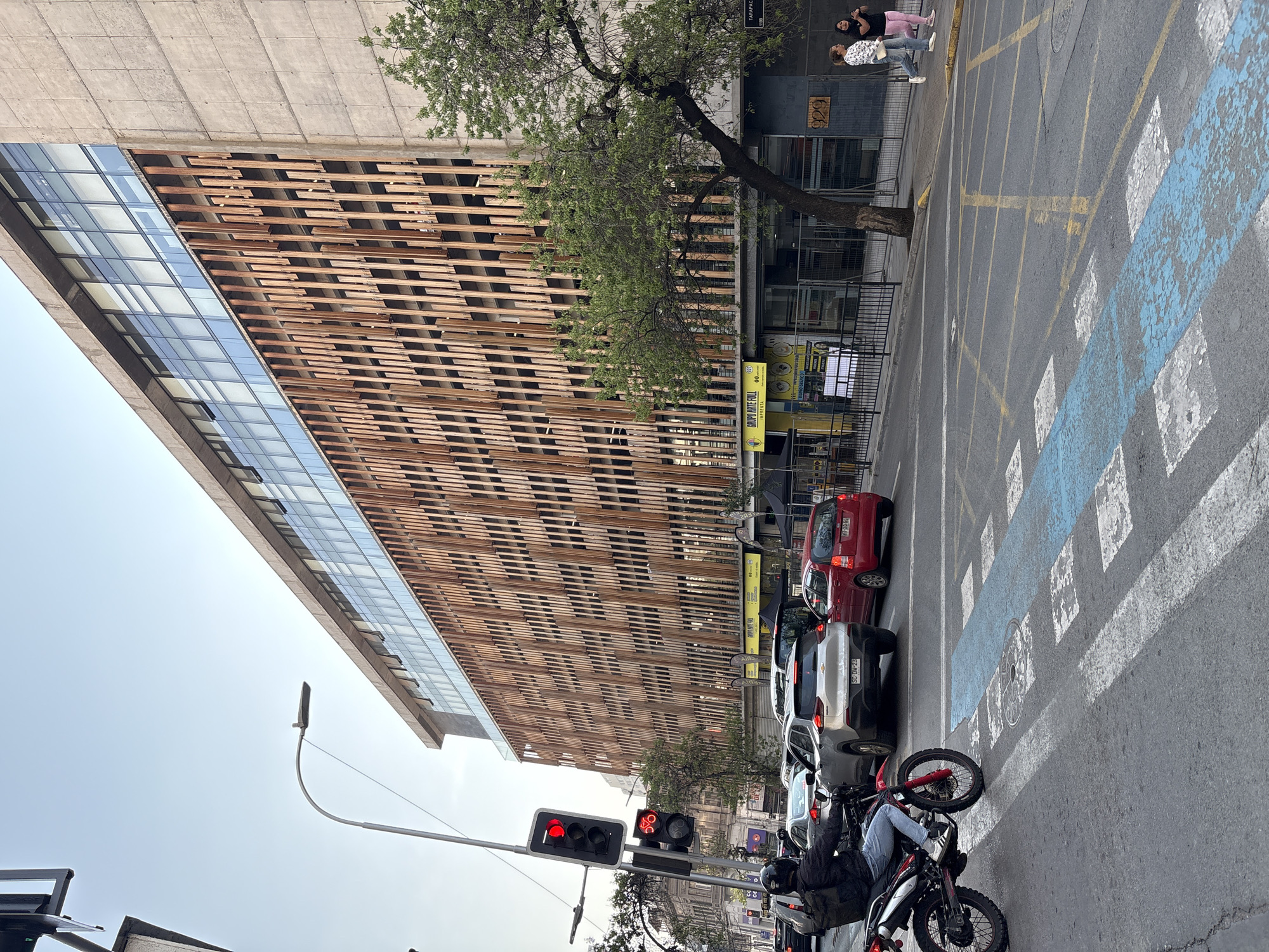 In Santiago, Chile, a busy street scene unfolds in front of a modern building with a distinctive wooden facade. Traffic is moderate, with several cars and a motorcycle visible on the asphalt road. A traffic light signals red for vehicular traffic, with a separate signal for bicycles illuminated. The building, a multi-story structure, features a repetitive pattern of vertical wooden slats over its lower levels, contrasting with smooth concrete on the upper portions and large glass windows. A mature tree with green foliage stands in front of the building. Two women walk on the sidewalk to the right of the frame. The street is marked with crosswalks, including blue and white painted pedestrian lanes, and yellow traffic lines. The sky is clear and overcast, suggesting a mild day.