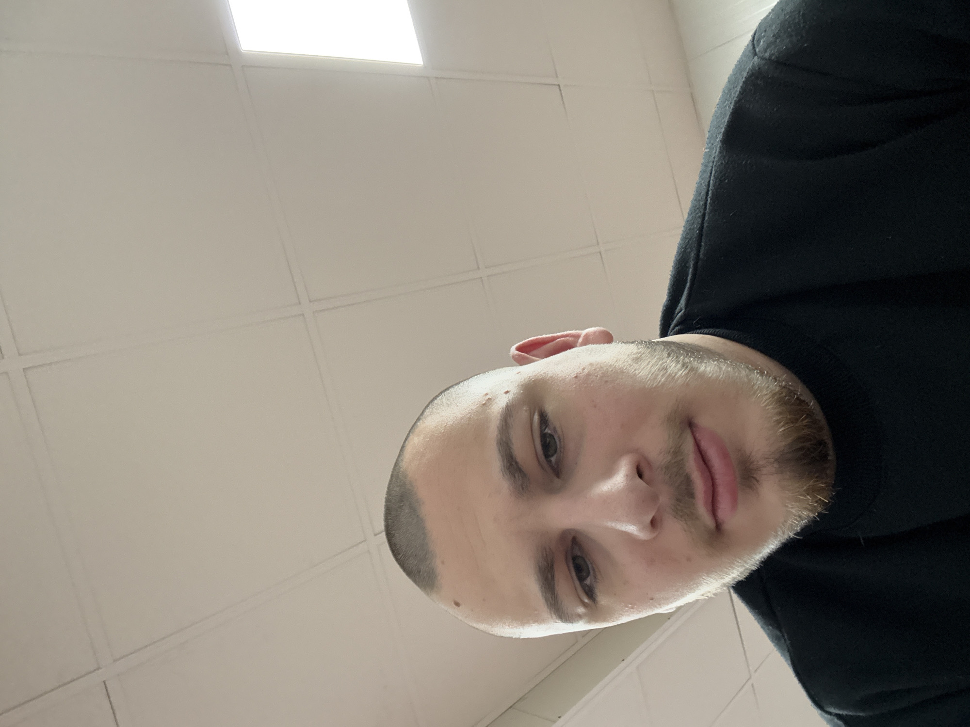 A man is shown in a low-angle selfie. He has a shaved head and a short beard. He is wearing a black sweatshirt. The background consists of a white, tiled ceiling with a fluorescent light panel. The man is facing the camera with a neutral expression. There is no information available to identify the location as Stara Zagora, Bulgaria.