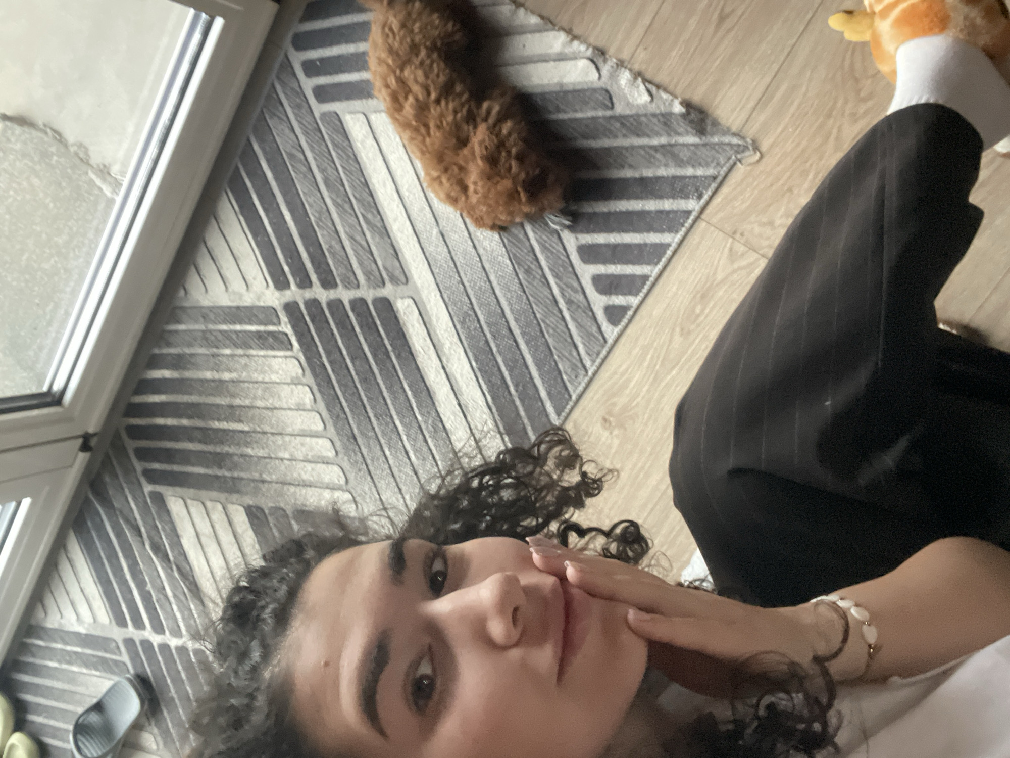 A woman with curly dark hair is lying on a light-colored wooden floor, resting her hand on her cheek. She is wearing dark pinstripe trousers and a white sock with a stuffed giraffe on it. A brown poodle is lying on a gray and white geometric patterned rug next to her. A window frame is visible to the left, and green sandals are near the window. The location is potentially an apartment in Sofia, Bulgaria.