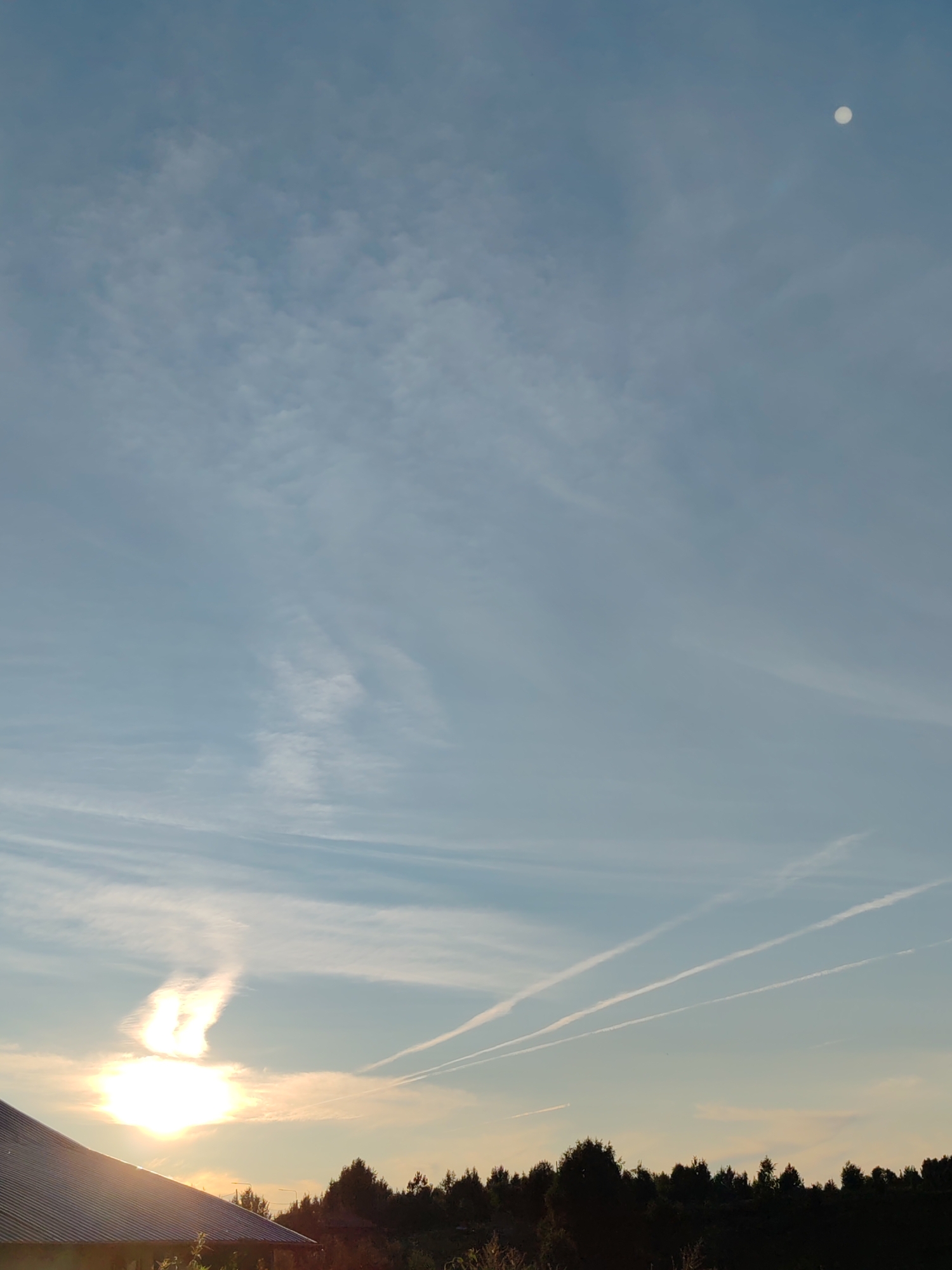 The image captures a clear, pale blue sky over a dark silhouette of trees and the rooftop of a building. In the upper right corner, the moon is visible as a bright white circle. Towards the lower left, the sun is a brilliant, partially obscured white sphere, casting a bright glow and creating an unusual, cloud-like formation above it. Several white contrails from aircraft streak across the sky at different angles. The overall scene suggests an outdoor, rural environment, possibly during sunrise or sunset, with a vast expanse of sky dominating the frame.