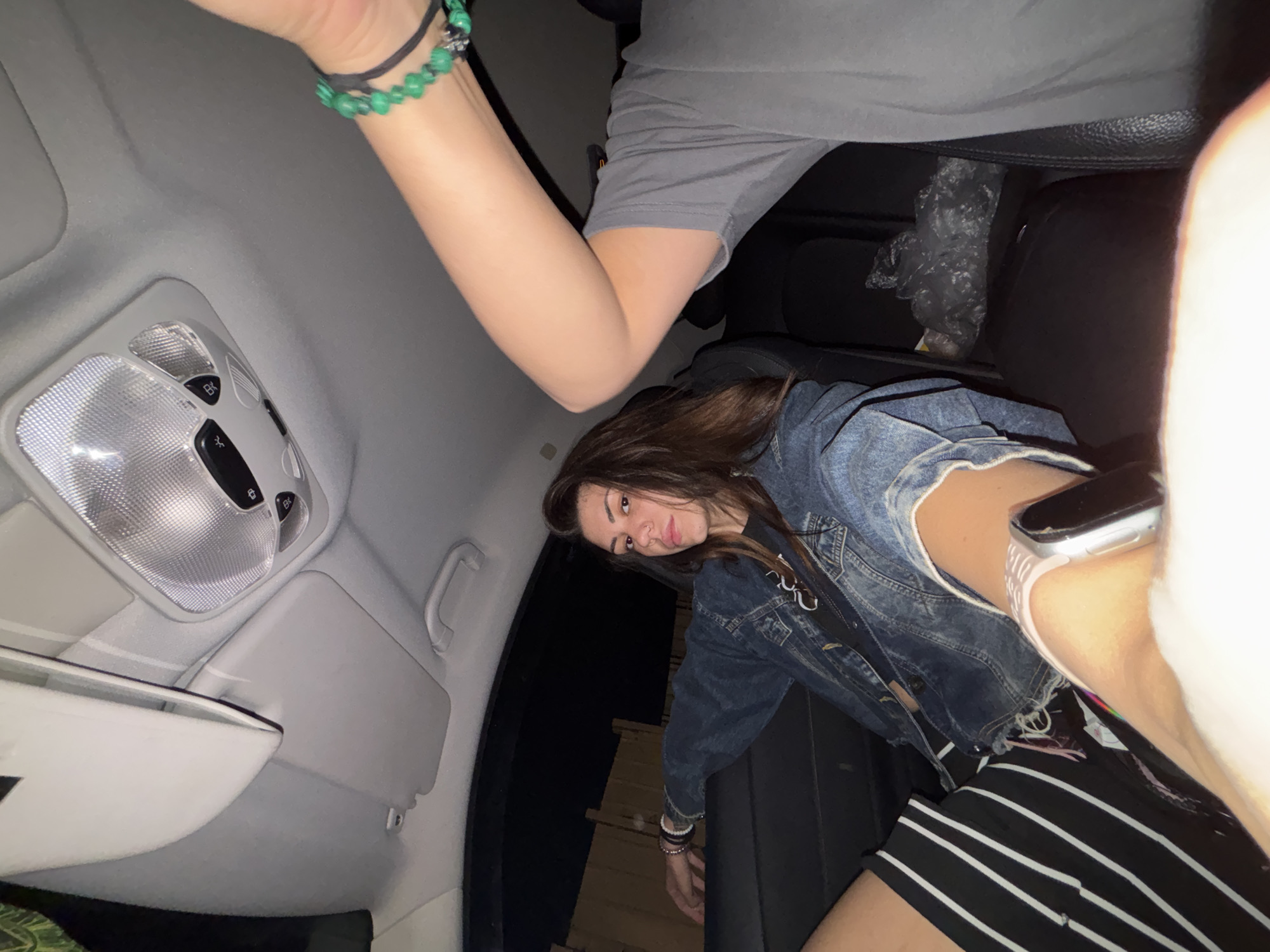 This image captures the interior of a car at night, located in the city of Sofia, Bulgaria. The scene is illuminated by interior vehicle lighting, creating shadows and highlights. A young female subject is prominently featured on the left side, seated in what appears to be the rear passenger seat. She has long brown hair, dark eyes, and a nose piercing on her right nostril. She is wearing a dark blue denim jacket over a black t-shirt, and black shorts or a skirt with white vertical stripes. An Apple Watch with a light-colored band is visible on her left wrist, and her left arm rests casually on the seat back. Her gaze is directed towards the camera, suggesting a posed portrait.
In the foreground on the right, the arm and upper torso of a second individual, likely the photographer, are partially visible. This person is wearing a grey t-shirt, and their right wrist displays multiple bracelets, including several with green beads and a black string bracelet. The low-angle perspective indicates the photo was taken from a position in front of the main subject, possibly by someone in the front passenger seat.
The car's interior features a light grey fabric ceiling with an overhead light console containing multiple clear lenses and control buttons. A grey sun visor is visible above the passenger seat area on the upper left, and a grey grab handle is on the upper right side of the ceiling. The car seats are dark, likely black or dark grey. Through the rear window, a dark background with what appears to be a wooden fence or structure is dimly visible, indicating the vehicle is parked or in an urban environment at night. A crumpled clear plastic bag rests on the seat or floor behind the main subject.