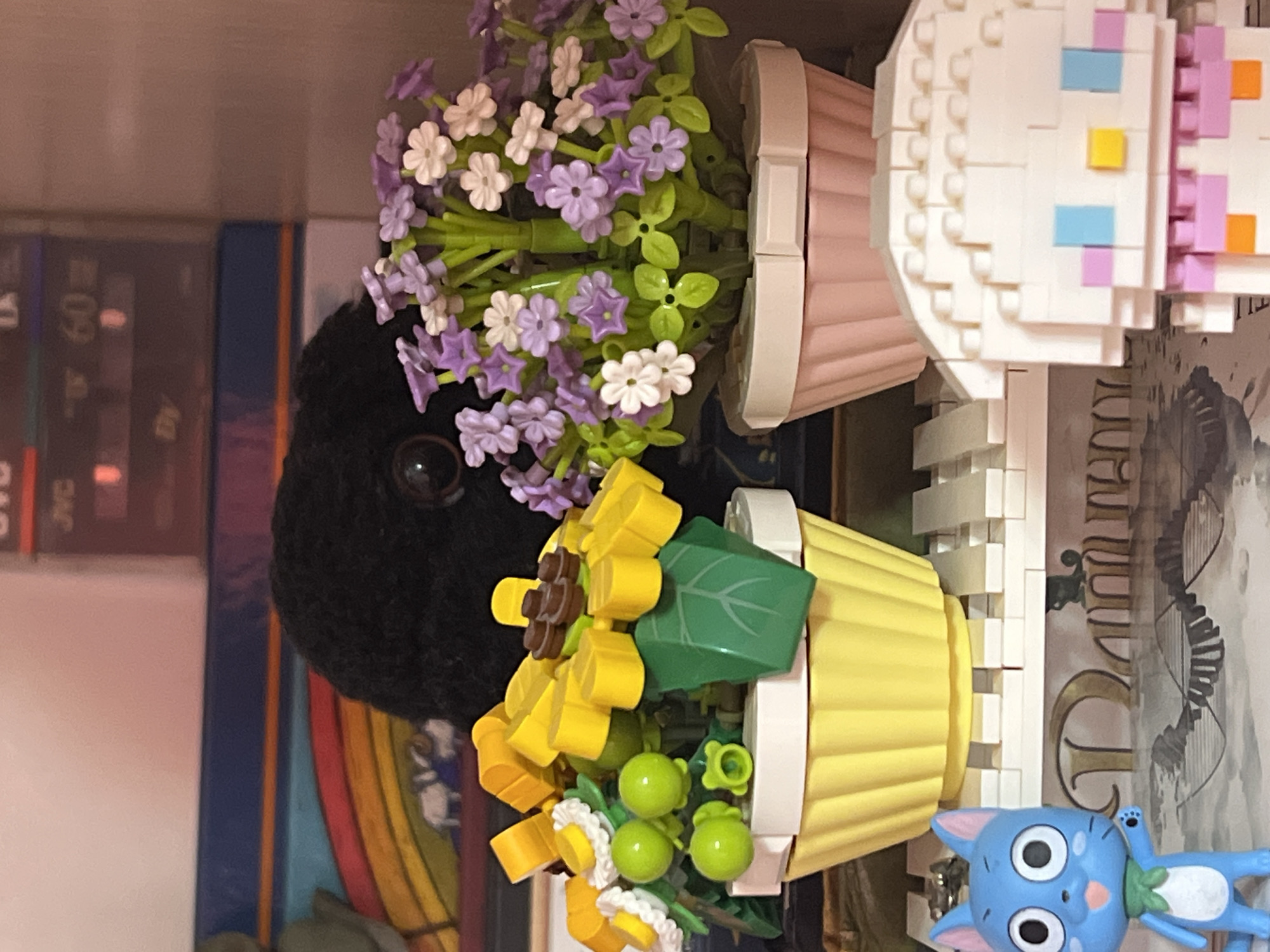 The image depicts a close-up indoor scene featuring Lego constructions and decorative items. In the foreground, to the left, is a blue cartoon cat figurine with large eyes and a green scarf. To the right is a Lego construction of a character with white hair, blue eyes, and pink and orange accents. Between these figures are two Lego potted plants. The left pot is yellow and contains yellow and white flowers with green berries. The right pot is pink and holds purple and white flowers with green leaves. Behind the plants, a black fuzzy object with visible eyes is partially obscuring the background. The background consists of shelves with various items, including what appears to be a vintage electronics device with a display showing "60". The scene is likely in a residential setting, possibly a shelf or display area, and is not associated with the city of Shymkent, Kazakhstan.