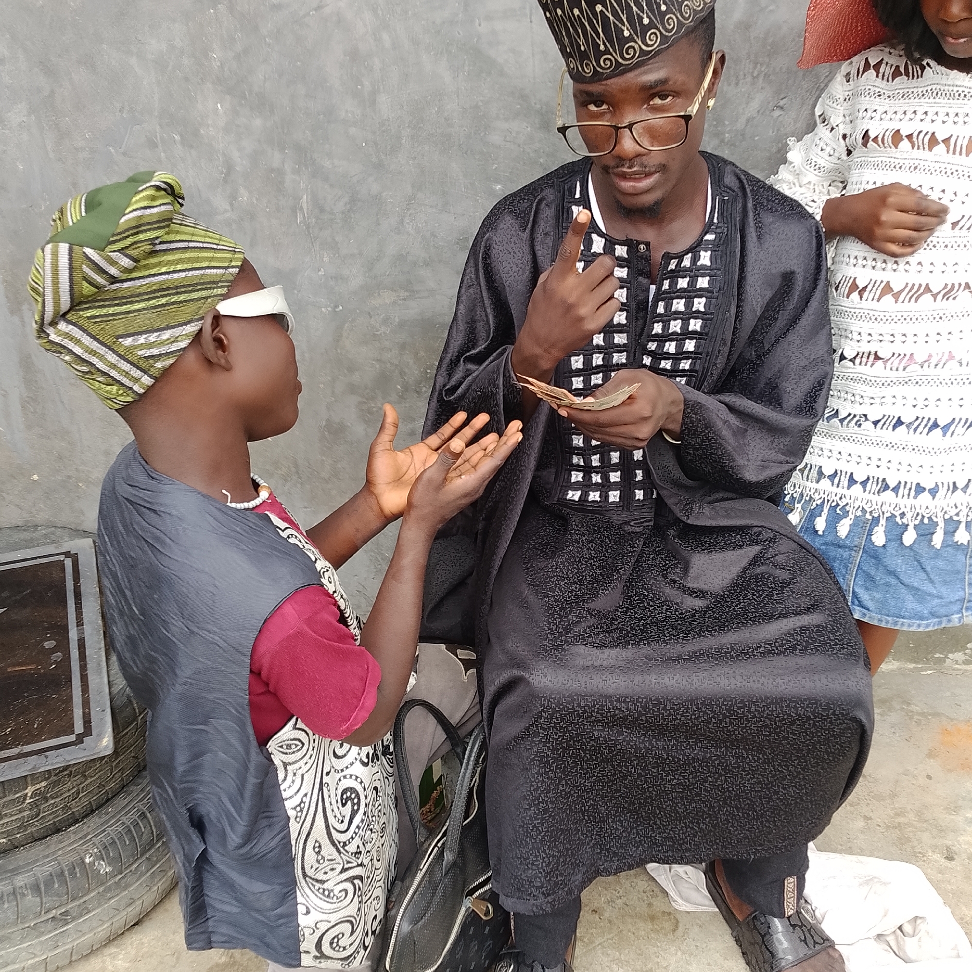 In the city of Ado, Nigeria, a man wearing a black traditional outfit and a patterned cap, along with glasses, is holding out a small stack of currency to another individual. The other person, wearing a green and white headwrap and a dark vest over a red shirt with a patterned lower garment, is reaching out with their hands. They are positioned outdoors against a gray concrete wall, with stacks of tires visible to the left and another person in a white outfit and a straw hat to the right.