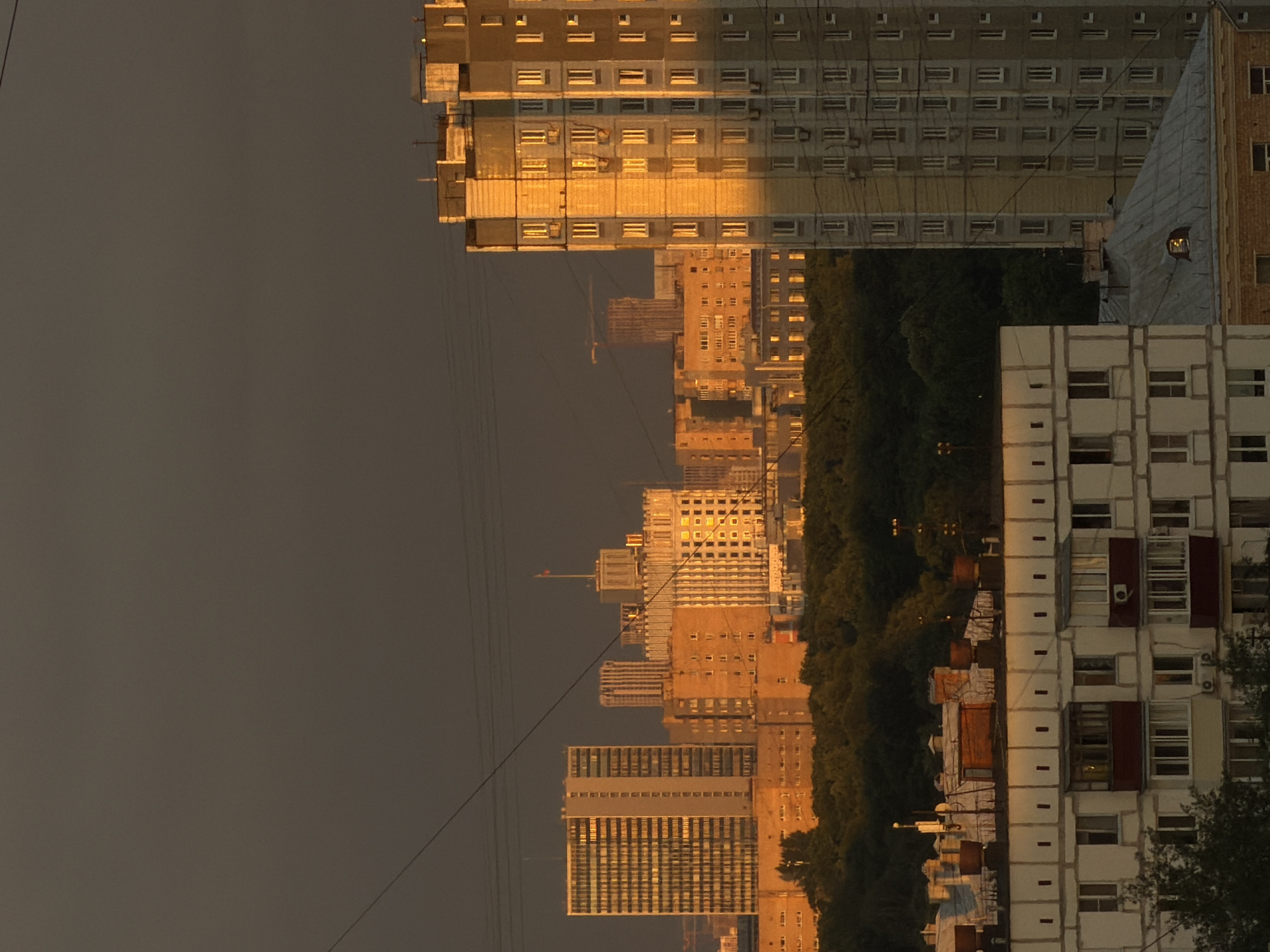 The image captures a city skyline at sunset, with buildings bathed in the warm, golden light of the setting sun. In the foreground, a multi-story apartment building with white facades and numerous windows is partially visible. Behind it, a dense forest stretches across the midground. Further in the background, a series of urban structures rise, including a prominent tall, light-colored apartment building on the right, illuminated by the golden light. Other buildings, varying in height and architectural style, are scattered throughout the cityscape, some appearing as older, brick structures and others as modern high-rises. The sky above is a dark, overcast gray. Power lines are visible crisscrossing the frame. The overall scene depicts a densely populated urban environment experiencing the dramatic lighting of sunset.
