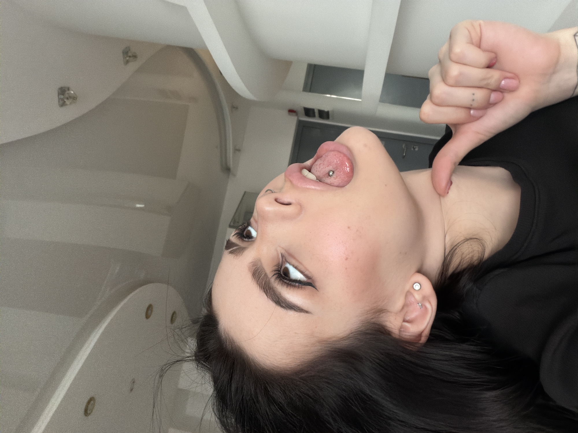 A close-up, portrait-style image, taken in Khimki, Russia, features a young woman with dark hair and striking dark eyebrows. Her head is tilted back, looking upwards and to the right, with her tongue extended, revealing a prominent silver tongue piercing. Her eyes are accentuated with dark eyeliner, and light freckles are visible on her cheek. She wears a silver stud earring in her left earlobe and a small silver cross-shaped earring higher on her ear. Her right hand is visible, making a "thumbs down" gesture; her fingernails are painted light pink, and a small tattoo of three dots is visible on her ring finger. She is wearing a black top with a scoop neck. The background consists of bright white, curved architectural features, possibly part of a ceiling or a large fixture, with several small, circular recessed lights embedded within them. The overall setting appears to be a modern indoor space, well-lit with artificial light, perhaps a spa or a contemporary bathroom.