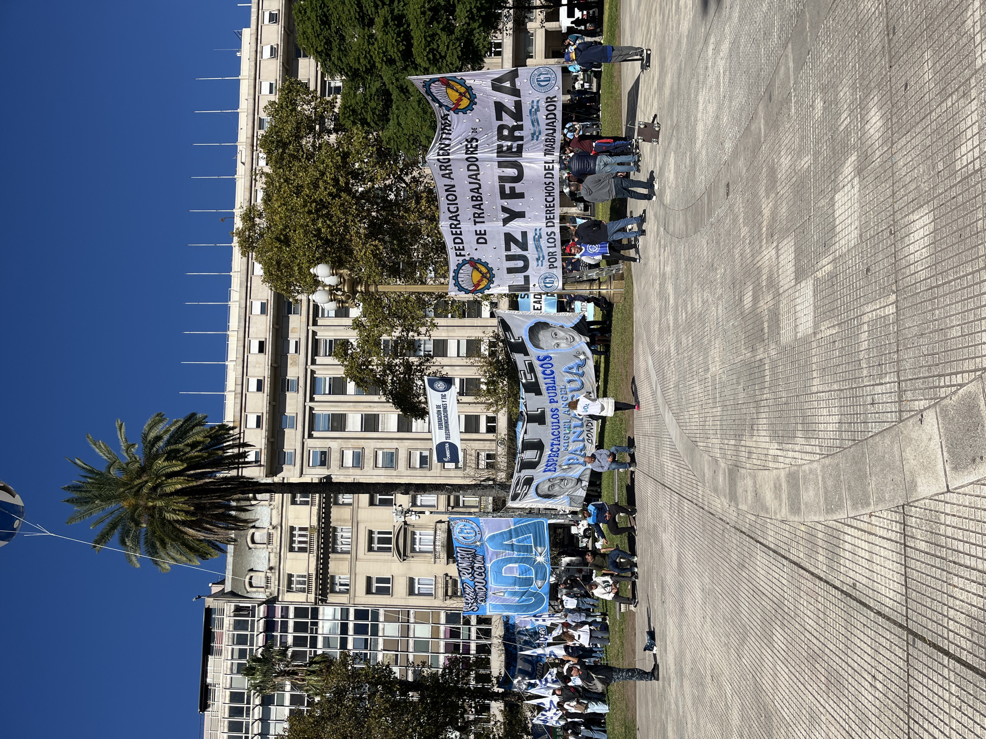 This image depicts an outdoor gathering or protest in a city square or park, likely in Buenos Aires, Argentina, under a bright, clear blue sky. The scene is dominated by a diverse group of people, primarily adults, many of whom are holding large banners and flags.
In the background, several multi-story buildings with numerous windows and balconies line the square, interspersed with lush green trees, including a prominent palm tree. The ground consists of extensive paved areas with a textured tile pattern, alongside patches of grass where some individuals are standing.
The central activity appears to be a labor union demonstration or assembly, characterized by the visible banners advocating for workers' rights. Notable text on these banners includes:
- "FEDERACION ARGENTINA DE TRABAJADORES DE LUZ Y FUERZA POR LOS DERECHOS DEL TRABAJADOR" (Argentine Federation of Workers of Light and Power - For the Rights of the Worker), accompanied by a logo with a gear and lightning bolt.
- A blue banner reading "SUTE ESPECTACULOS PUBLICOS MIGUEL ANGEL CONDUCCION," featuring a portrait of a man.
- Other banners mention "FEDERACIÓN DE FoesITRA TELECOMUNICACIONES Y TIC" (Federation of Foesitra Telecommunications and ICT) and "SEÑO ROMERO CONDUCCION" (Teacher Romero Leadership), with "UGA" also visible.
The event takes place on a sunny day, with the light casting shadows on the paved ground. The overall impression is that of an organized public event in an urban setting.
