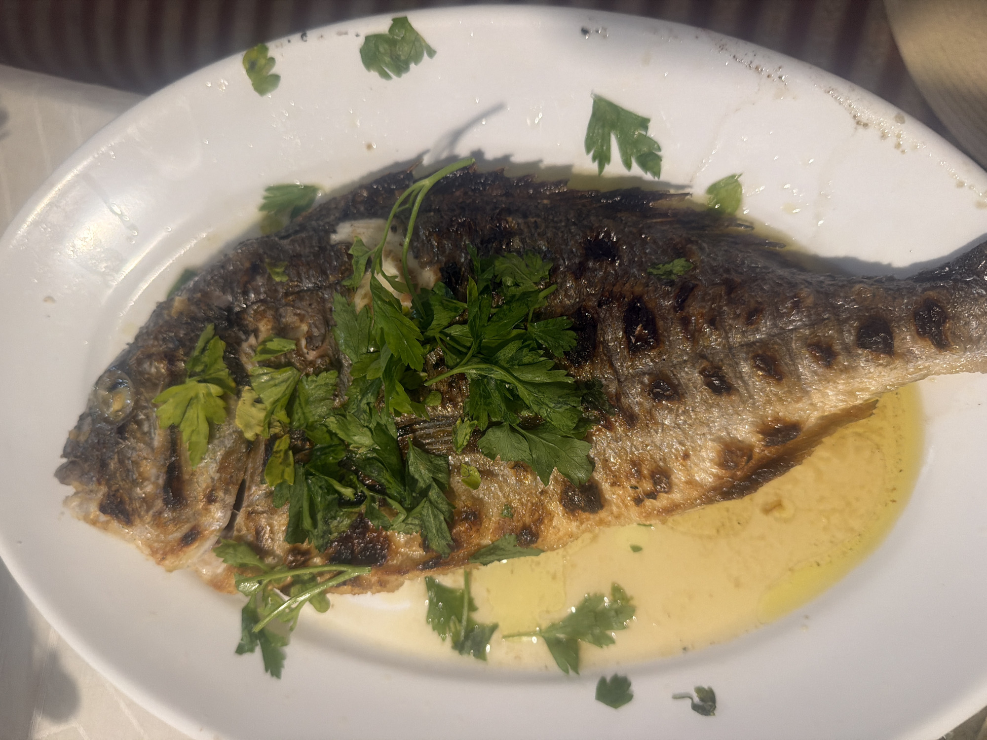 An overhead shot shows a whole grilled fish garnished with chopped parsley, served on a white oval plate. The fish, which appears to be a sea bream or similar, is golden brown with visible grill marks and has a slightly charred texture. The parsley is sprinkled generously over the top, with some sprigs also scattered around the fish in the yellowish liquid that coats the bottom of the plate. The plate is set on a textured surface, possibly a tablecloth. The background is blurred, suggesting an indoor dining setting. The image does not contain any information about its location in the city of Mandra, Greece.