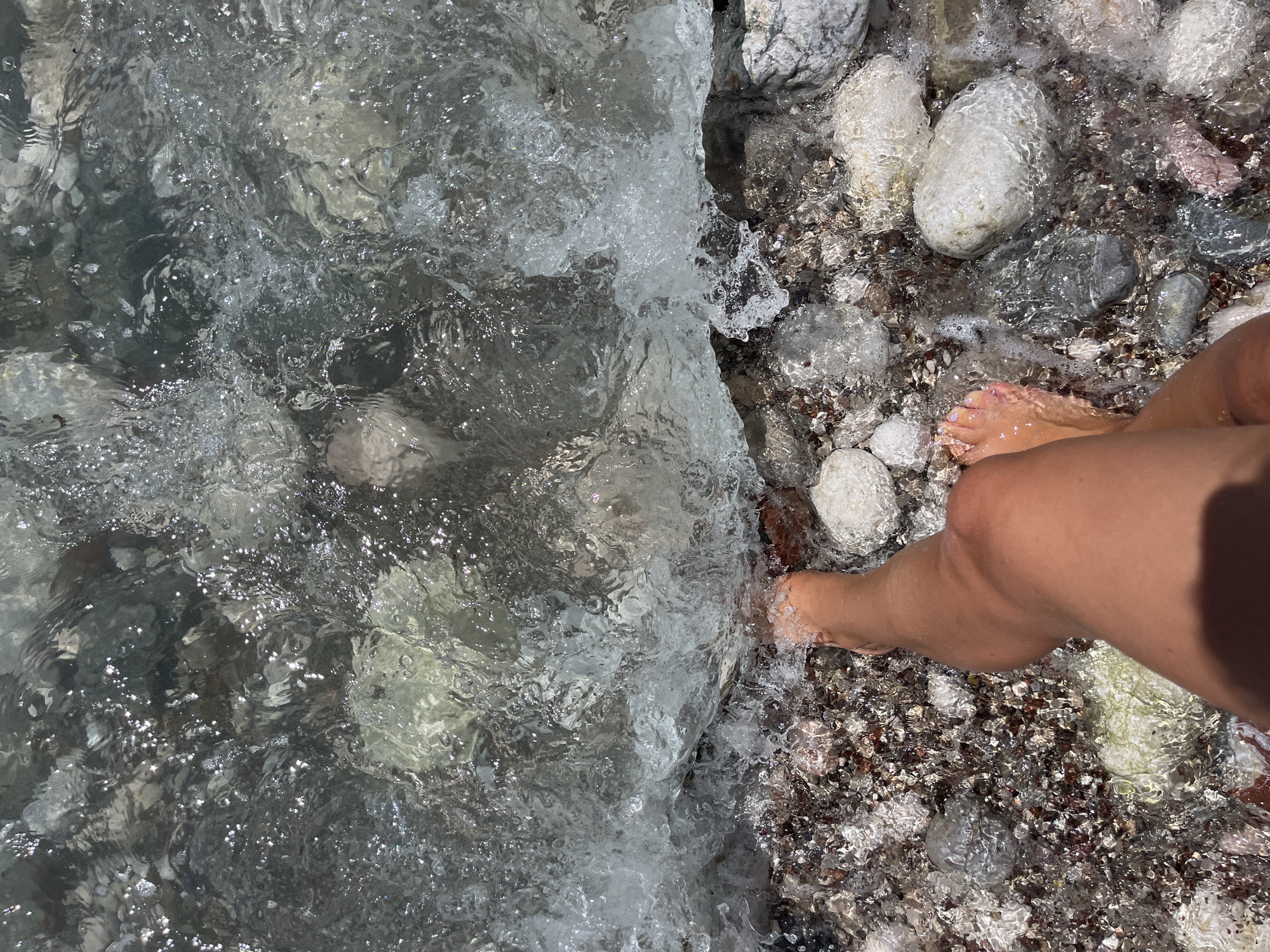 The image shows a close-up view of a person's bare legs and feet standing in shallow, clear water on a rocky beach. The water is gently lapping at the shore, with small waves and white foam. The seabed is covered with various sizes of smooth, grey, white, and reddish-brown pebbles, some of which are partially submerged. The person's skin tone is tan, and their toenails appear to be painted with a light pink or purple polish. The scene suggests a relaxing moment at the seaside, possibly in the city of Bar, Montenegro, known for its coastline with pebble beaches.