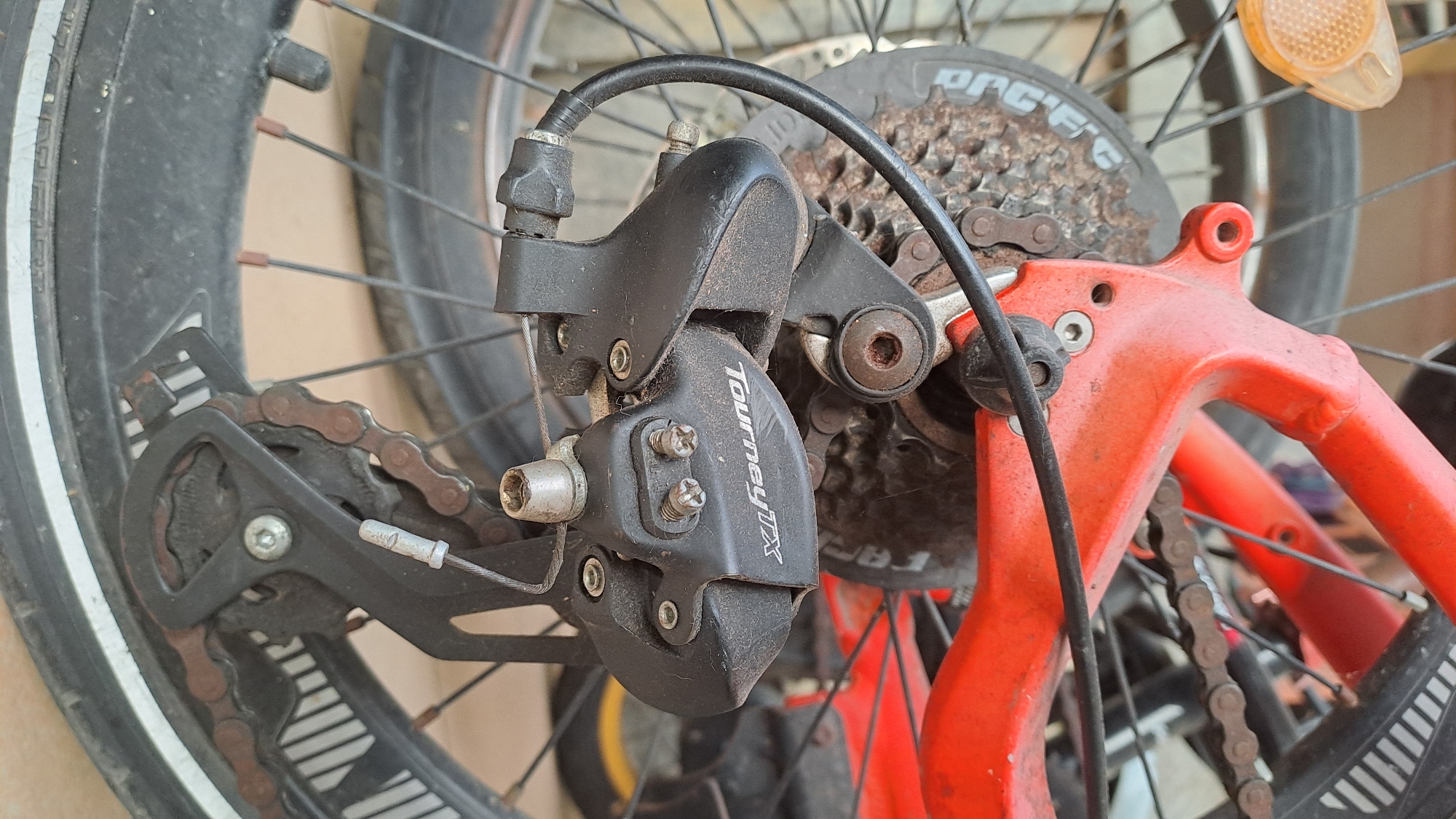 The image displays a close-up view of a bicycle's rear derailleur and cassette. The derailleur is black and has "Tourney TX" branding visible. The cassette, located behind the derailleur, appears to be made of metal and shows signs of rust. A rusty bicycle chain is visible, running over the cassette and through the derailleur. The bicycle frame is painted red, and parts of a black bicycle wheel are visible in the foreground and background. The overall scene suggests an outdoor setting, possibly in a city like Lhokaweawe, Indonesia, given the mention. There are no people or animals present in the image. The lighting is natural, and the focus is on the mechanical components of the bicycle.