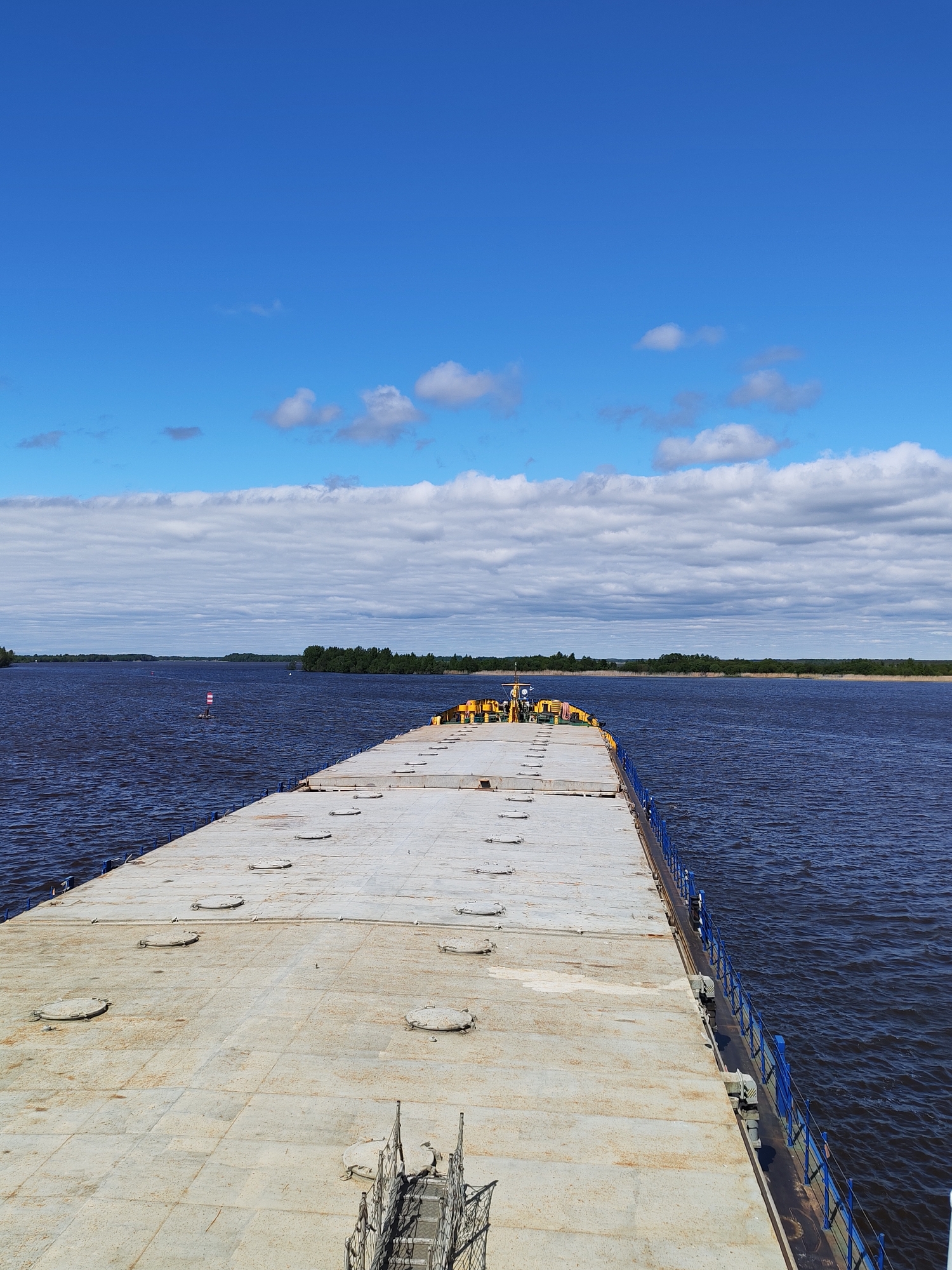 The image shows a large, flat-topped barge on a river in Kosino, Russia. The barge's deck is composed of large, rectangular sections with circular hatches. A blue metal railing runs along the edge of the barge on the right side. In the distance, there's an island with trees on it. A navigation marker can be seen in the water, slightly to the left of the barge's center. The sky is blue with scattered clouds. The river's water is dark. There are people and machinery located on the barge, towards the background of the shot.