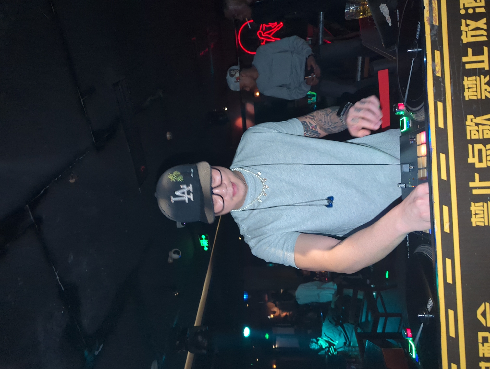 A man in a light grey t-shirt and baseball cap is operating a DJ mixer with a turntable in what appears to be a dimly lit club. The mixer is decorated with yellow stripes and Chinese characters. There are other people in the background, some sitting at tables, and neon lights are visible. While I do not have enough information to confirm the exact location, the presence of Chinese characters suggests it might be in China, and given the prompt, possibly in the Liangjiang area.