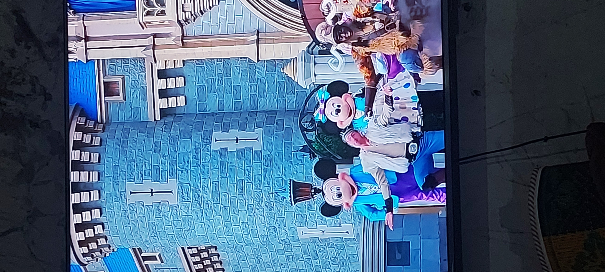 The media file displays a television screen showing a scene with Disney characters and performers. In the foreground, two individuals in Mickey Mouse costumes are visible. One of them is in a blue outfit and the other is in a teal and purple outfit, with Minnie Mouse also present in a pink and teal outfit. They appear to be part of a performance or parade, possibly at a theme park. Behind the characters, a large, blue castle-like structure is visible, resembling Cinderella Castle at Walt Disney World. The castle is adorned with windows and architectural details, and the overall setting suggests a magical or celebratory event. The lighting appears to be bright, indicating daytime. The image is captured from an angle, with a dark, textured surface in the foreground and background, possibly indicating the viewer's surroundings. No visible text is present in the image. The context of South Tangerang, Indonesia, suggests this footage might be from a broadcast or a captured moment within that region, but the content itself is universally recognizable as a Disney-themed event.