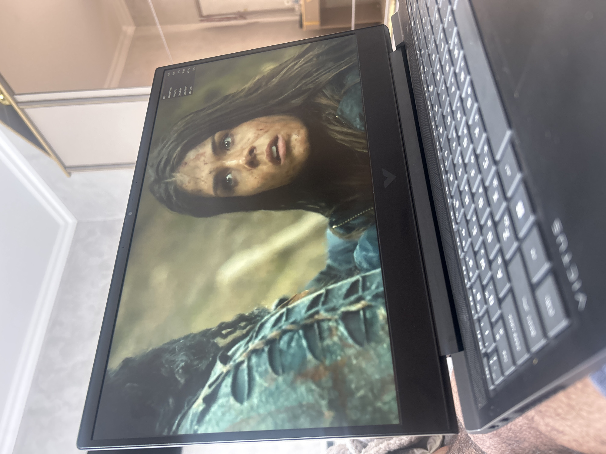 A close-up, angled shot shows a black laptop with the screen displaying a still image of a woman with dirty skin, wide eyes, and slightly open mouth. The image suggests she is in a state of distress or shock. The background of the screen image is blurry and appears to be a forest or natural setting. The laptop's keyboard is visible in the foreground. The laptop appears to be an HP Victus laptop. The top left corner of the screen also displays performance metrics like FPS, Frame Time, GPU Util, GPU Power, GPU Temp (42°C), and CPU Util. The location Topolovgrad, Bulgaria is not relevant as it cannot be determined from the image alone.