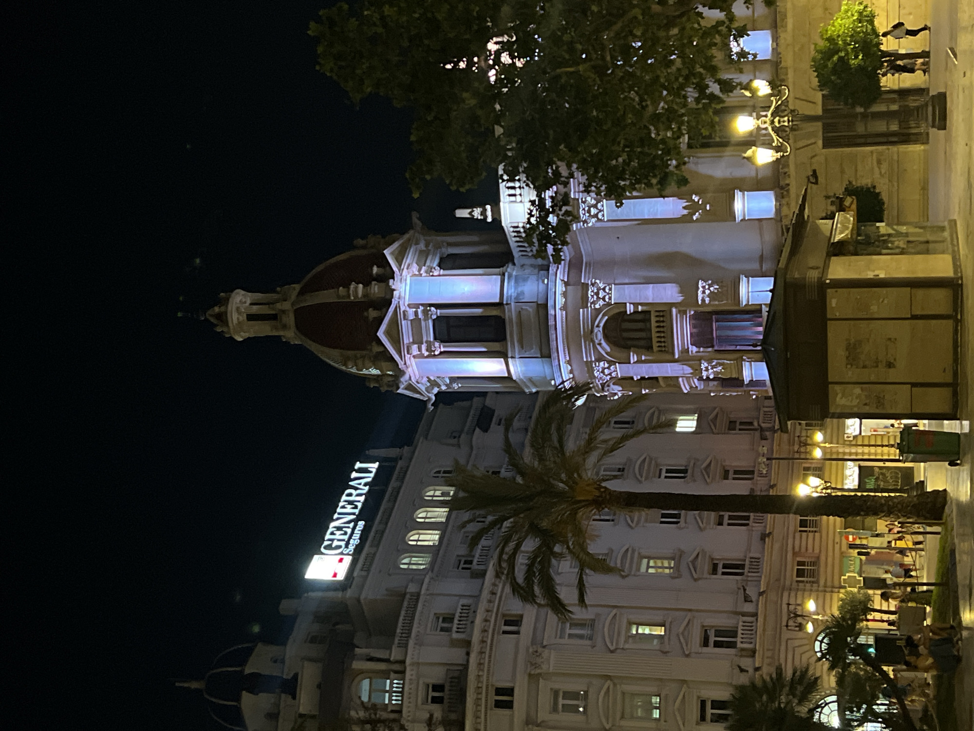 At night in València, Spain, a tall, light-colored building with a domed roof is illuminated. A "GENERALI Seguros" sign is visible. A palm tree stands in front of the building. People are walking on the sidewalk in the foreground. A newsstand is present. Streetlights provide additional illumination.