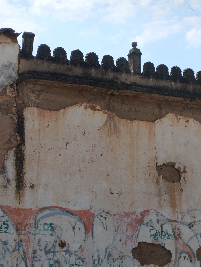 The image depicts a deteriorated building wall in Guacara, Venezuela, viewed from a low angle against a partially cloudy, light blue sky. The upper section of the wall features a dark, ornate parapet with a series of repeating decorative elements, including two distinct finials: a rectangular column on the left and a spherical top on a column towards the right. Below the parapet, the wall's light-colored plaster or render is extensively damaged, with large, irregular sections missing, revealing the darker, reddish-brown construction material underneath. Dark vertical streaking, suggestive of water damage, is visible on the left side of the wall, and a faint radial stain pattern is present in the upper-middle portion of the exposed substrate. The lower third of the wall is covered in multiple layers of graffiti, including large, light-colored bubble letters outlined in dark tones over a faded orange-red band, overlaid with smaller, darker green and black spray-painted text and tags. Several holes in the plaster are evident within the graffiti-covered area, exposing the underlying material. No individuals, animals, or discernible activities are present in the scene.