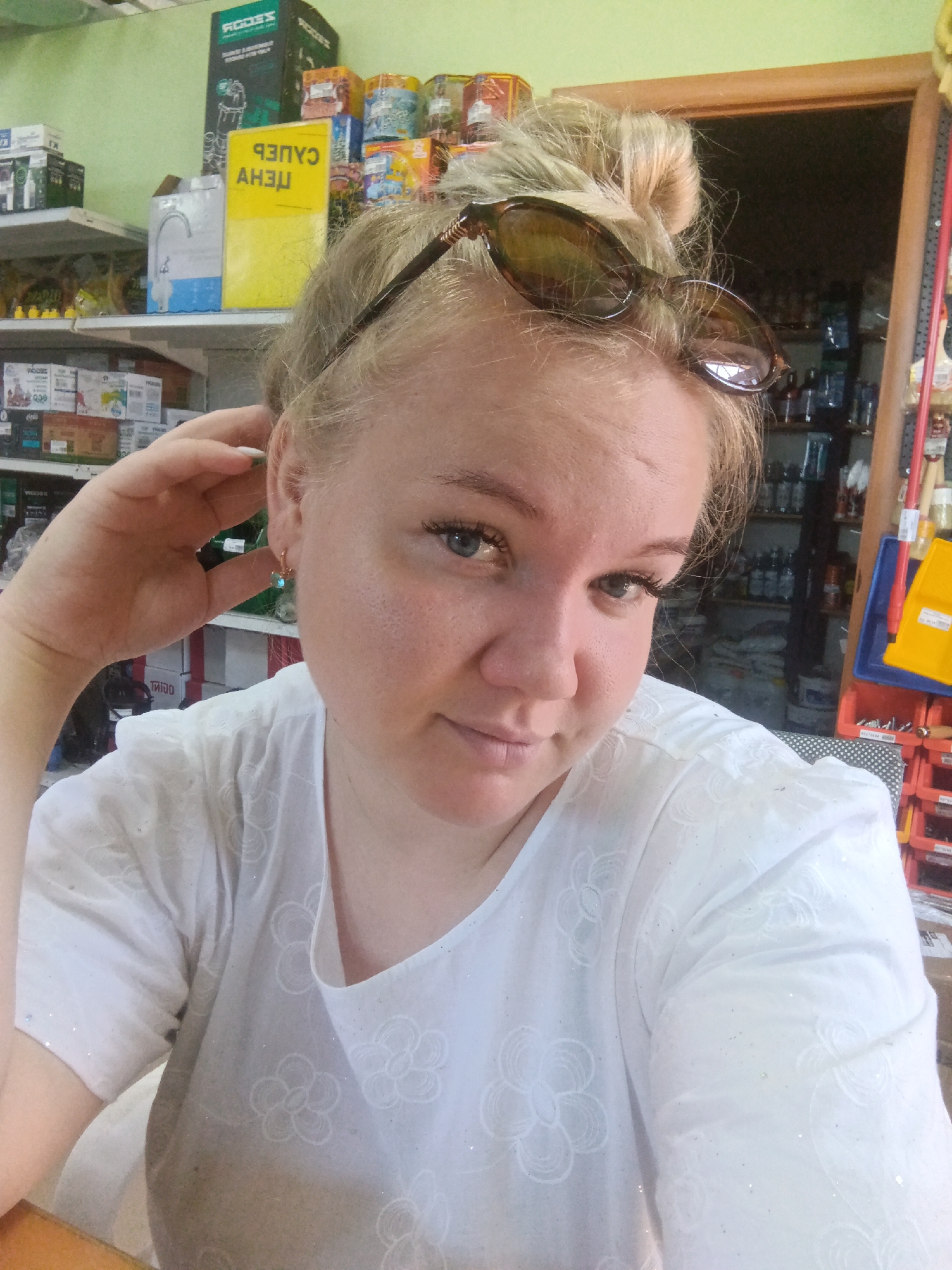 A light-skinned woman with blonde hair styled in a bun is looking at the camera. She is wearing a white shirt with a floral pattern. Her right hand is raised to her ear. She is wearing sunglasses on top of her head. The background appears to be an interior of a store with shelves stocked with various items, including boxes and bottles. There is also a door frame visible. The setting is likely a retail environment, possibly a hardware or general store.