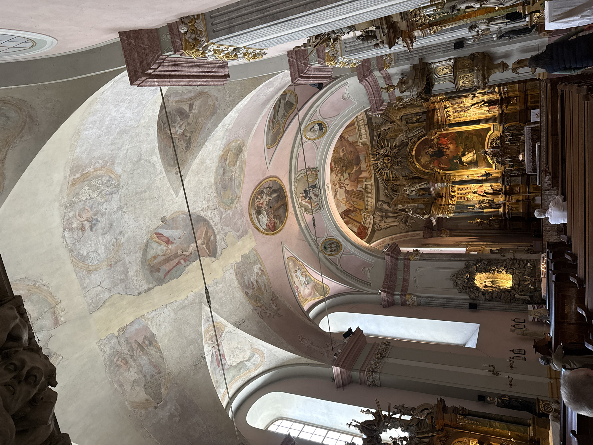 This image depicts the interior of the Saint Anne Church in Budapest, Hungary. The view is angled upwards, capturing the ornate, fresco-covered ceiling. The frescoes depict religious scenes and figures, rendered in faded colors with noticeable cracking and aging. Decorative arches and moldings frame the frescoes, adding to the church's elaborate baroque style. Below the ceiling, windows provide natural light, illuminating the interior. Carved wooden elements, including statues and decorative panels, are visible along the walls and in the sanctuary. People are present, some sitting in pews, and others standing near the altar area. The overall scene conveys the grandeur and historical significance of the church.