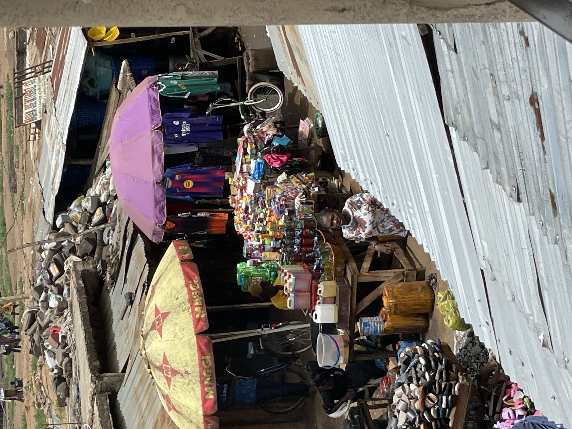 This photograph, taken in Bukuru, Nigeria, presents a scene of a marketplace. The view is oriented such that objects appear rotated.
In the foreground, sections of corrugated metal roofing are visible. Several small shops are located in the foreground. The shops feature open-air displays of goods. Items seen on display include clothes, a variety of bottled and packaged goods, and footwear. Shop umbrellas, one featuring a "Maggi" logo, provide shade. Bicycles are also present.
People are interspersed among the shops. Some of these people appear to be tending to the displays and others seem to be customers.
In the background, there is a view of other buildings and structures, as well as what appears to be vegetation and open space.