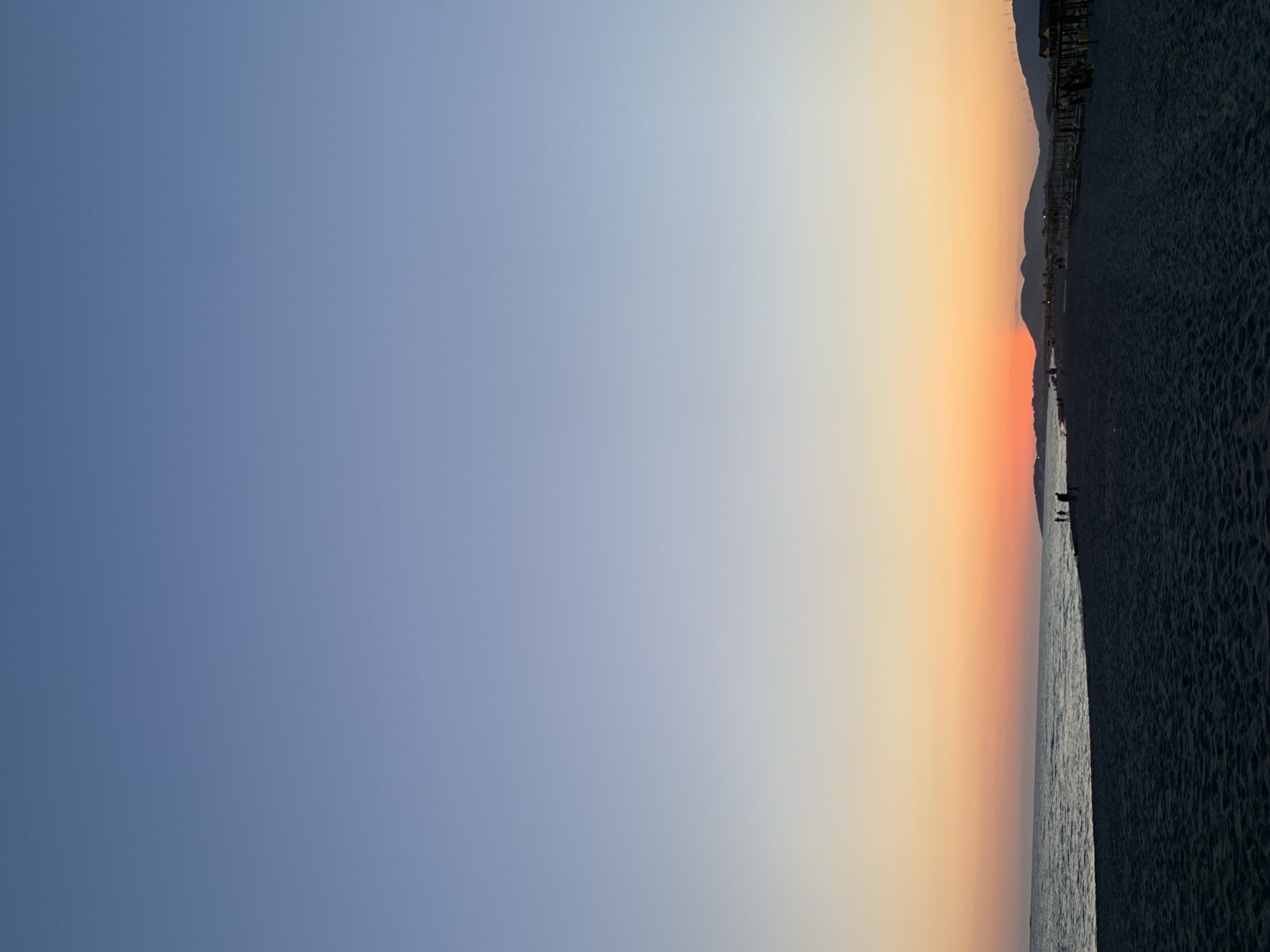 The image depicts a tranquil beach scene at sunset in Donji Štoj - Shtoji i Poshtëm, Montenegro. The sky transitions from a clear, pale blue at the top to warm hues of orange and pink near the horizon, where the sun is setting. A calm sea with gentle waves meets a dark, sandy beach. Silhouettes of a few people are visible on the beach and near the water's edge, suggesting a quiet evening. Along the coastline, there are rows of beach umbrellas and loungers, indicating a developed resort area. In the background, distant hills and wind turbines complete the picturesque landscape. The overall atmosphere is peaceful and serene, characteristic of a coastal evening.
