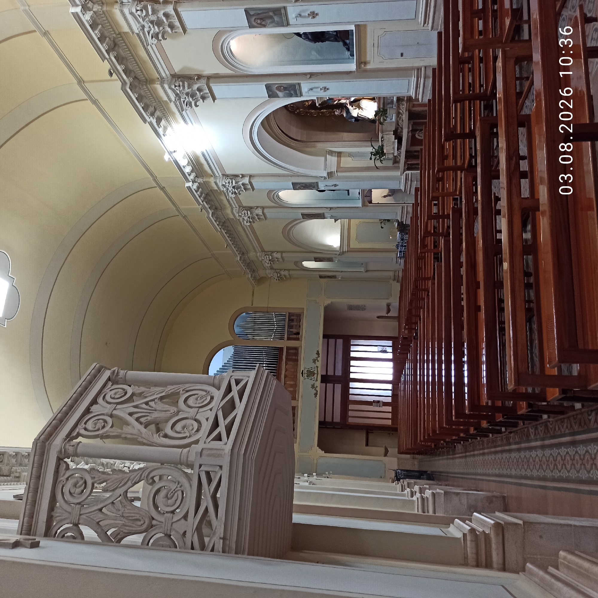The image shows the interior of the Chiesa Matrice, also known as the Duomo di Ruvo, located in Ruvo di Puglia, Italy. The perspective is from the nave, looking towards the altar. Rows of polished wooden pews fill the central space of the church. To the left, a ornately carved pulpit juts out from a pillar. Further down the nave, large wooden doors are visible, with light streaming through their panels. The walls are adorned with classical architectural elements, including columns and arches, with niches containing religious statues and paintings. An organ is visible in the upper section of the left wall. The ceiling is a pale yellow, with arched sections and decorative trim. The floor is tiled with a patterned runner extending down the center aisle. The date stamp in the lower right corner indicates 03.08.2026 10:36. No people are visible in the scene.