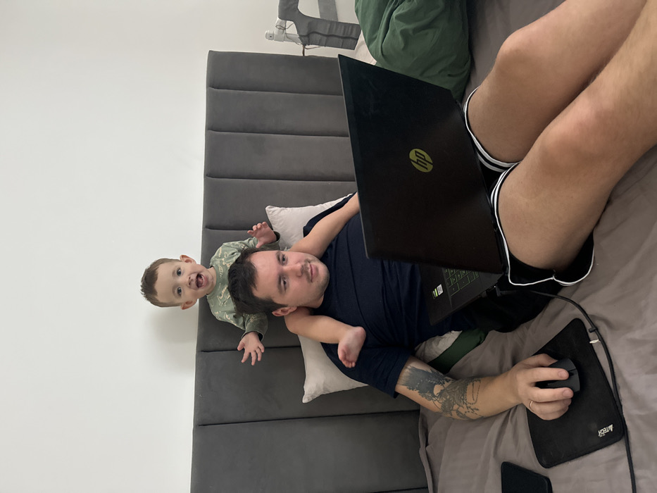 An adult male and a young child are depicted indoors in what appears to be a bedroom in Селекционер-2 Снт, Russia. The adult male, with short dark hair and a tattoo on his left forearm, wears a dark blue t-shirt and black shorts with white trim. He is reclining on a bed with grey bedding, operating a black HP laptop positioned on his lap, and a black wired mouse on a black mousepad to his left. A dark smartphone rests next to the mousepad. A light-colored pillow supports his head against a dark grey upholstered headboard with vertical panels. A male toddler, with light brown hair and wearing a green long-sleeved shirt with a pattern, is seated on the adult's shoulders, with his legs resting on the adult's upper back. The child has his arms raised and spread slightly, and his mouth is open in an expression of excitement. The background consists of a plain white wall. To the right, a dark green fabric item and a white electronic device are partially visible.