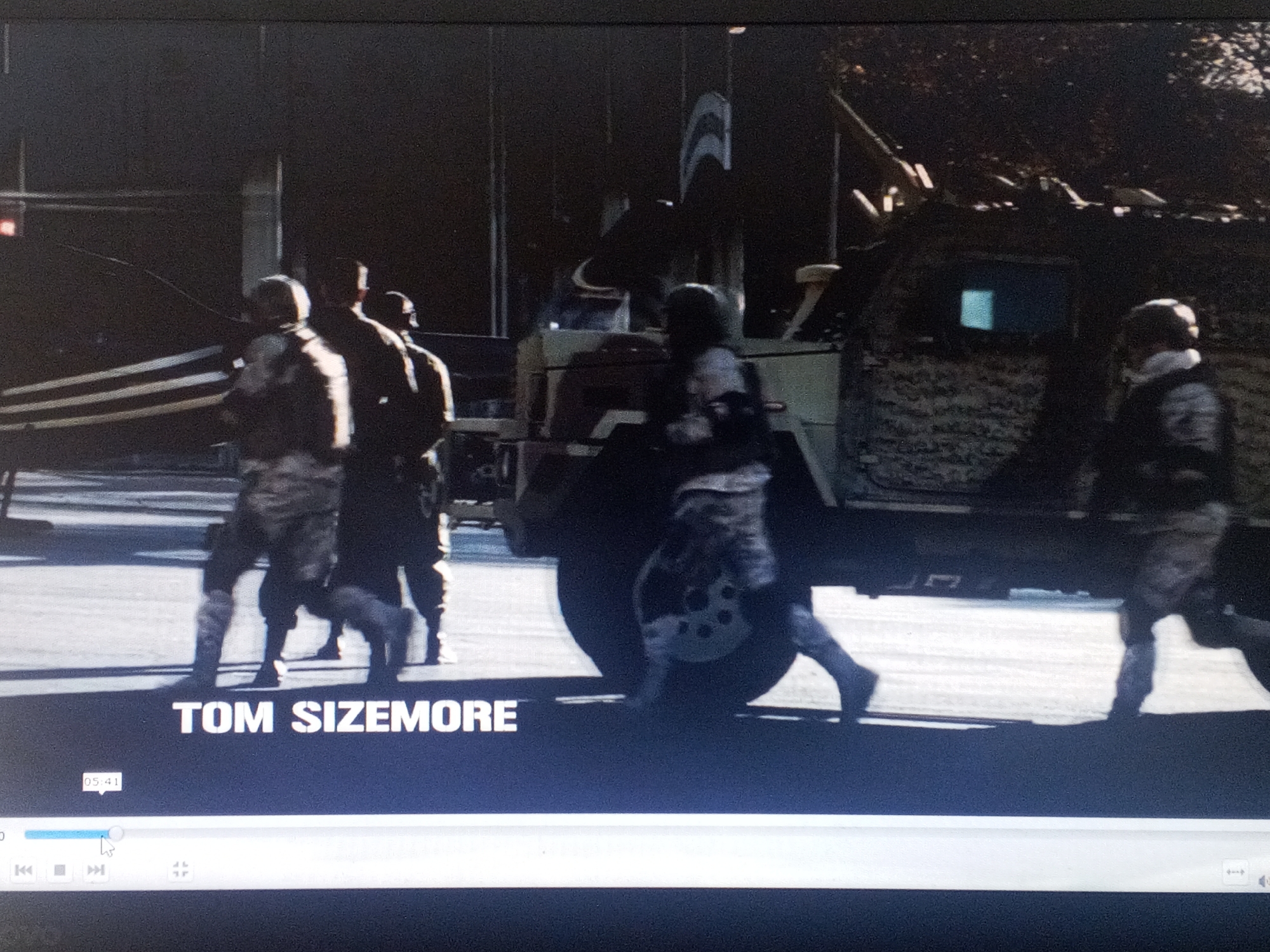 The image depicts a scene with several people walking on a street in an urban environment. The name "TOM SIZEMORE" is displayed at the bottom of the frame. An armored vehicle, partially covered in camouflage or netting, is positioned to the right of the group. There is a flagpole-like object with a curved design on top, attached to the vehicle. Other figures are partially visible to the left of the vehicle, and the overall lighting suggests an outdoor, daytime setting. The precise location is unknown.