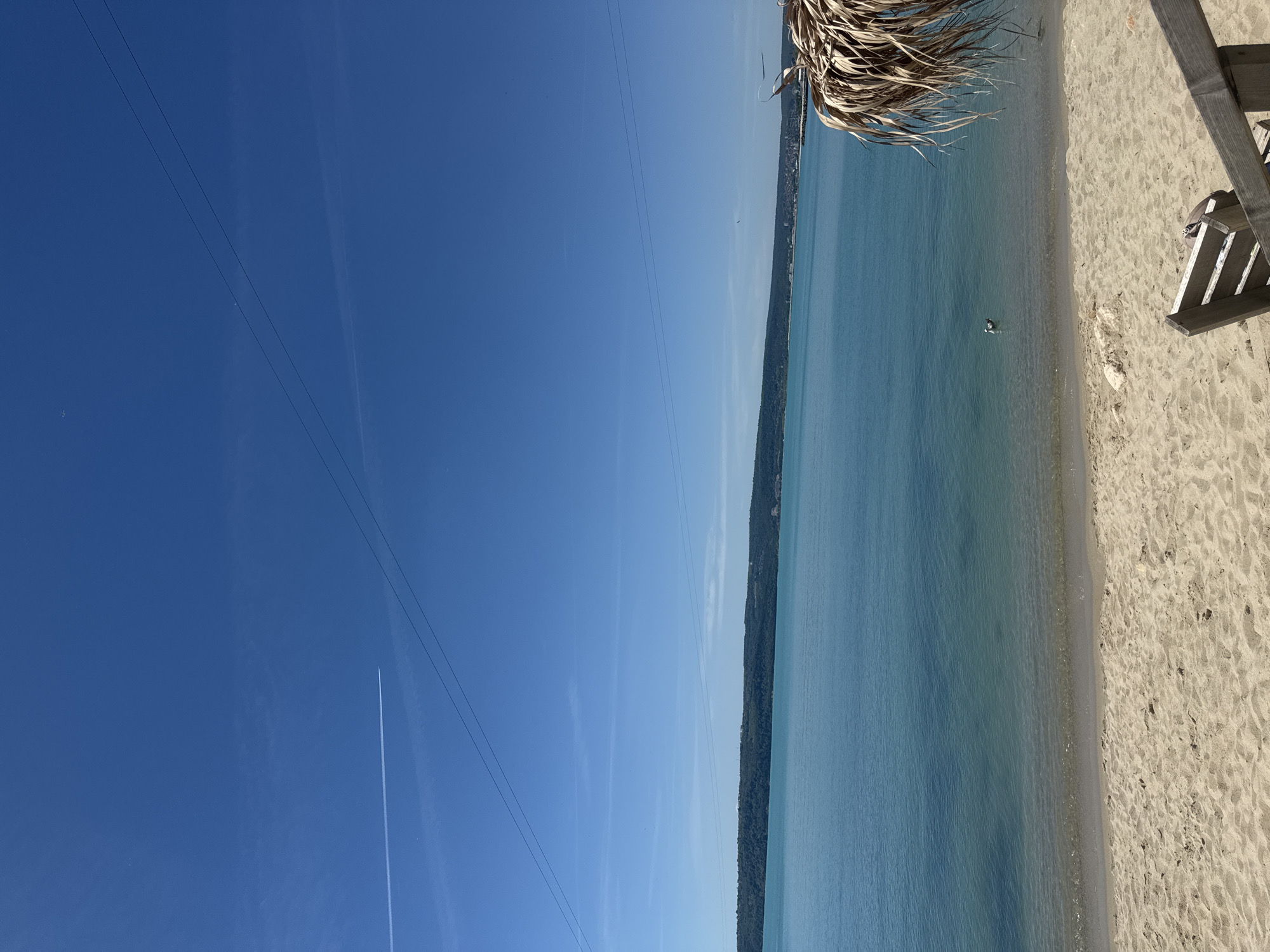 The image shows a view of a beach scene in Varna, Bulgaria. The sky is a clear blue with some power lines. The sea is turquoise and there is a thin strip of land covered with trees visible on the other side of the water. The beach is sandy and there is a part of a thatched umbrella and a beach chair in the frame. There appears to be a bird standing in the shallow water.