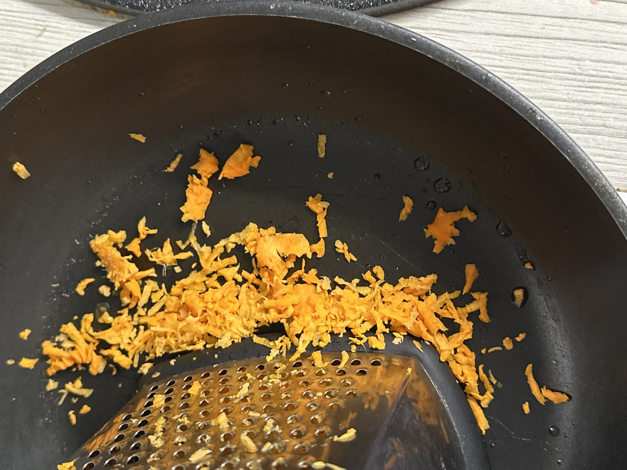 A close-up shot of a black pan containing shredded orange material, likely carrot or other root vegetable. A grater is visible at the bottom of the pan, suggesting the material was freshly grated. The pan and grater are resting on a light-colored wooden surface. There is no indication of the image being located in Chaikovsky, Russia. There are no people visible in the image.