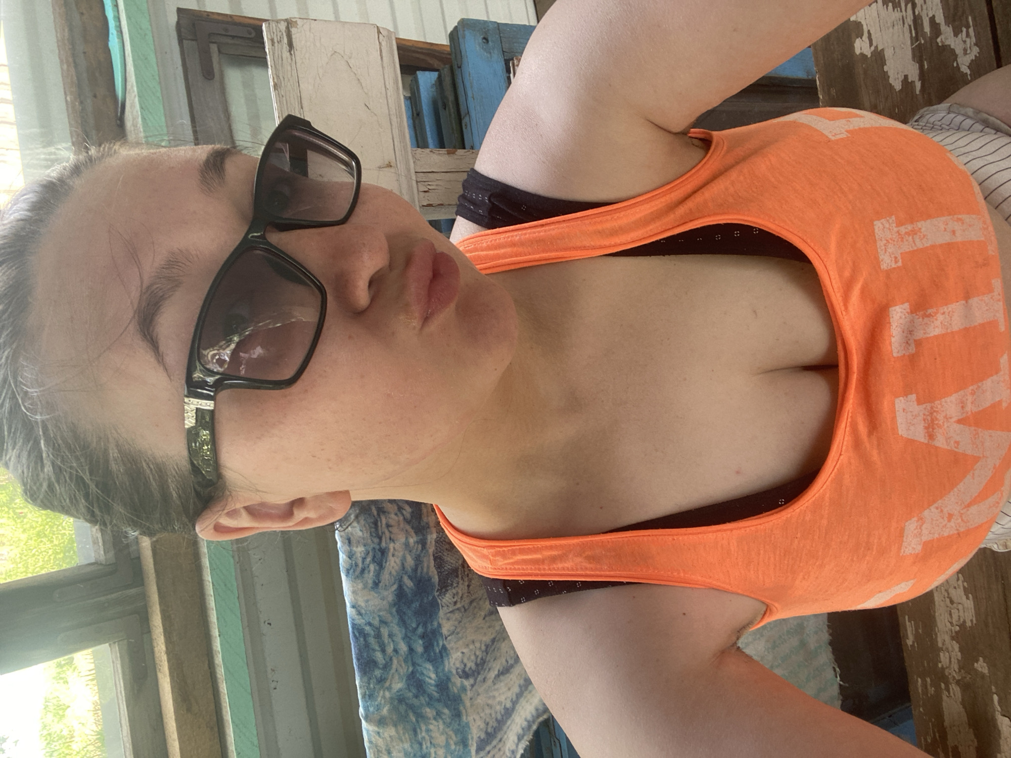 A woman with sunglasses and an orange tank top is photographed from above. She is seated in a location with weathered wooden surfaces and a window. The specific location within the city of Прикамский, Russia cannot be determined from the image.