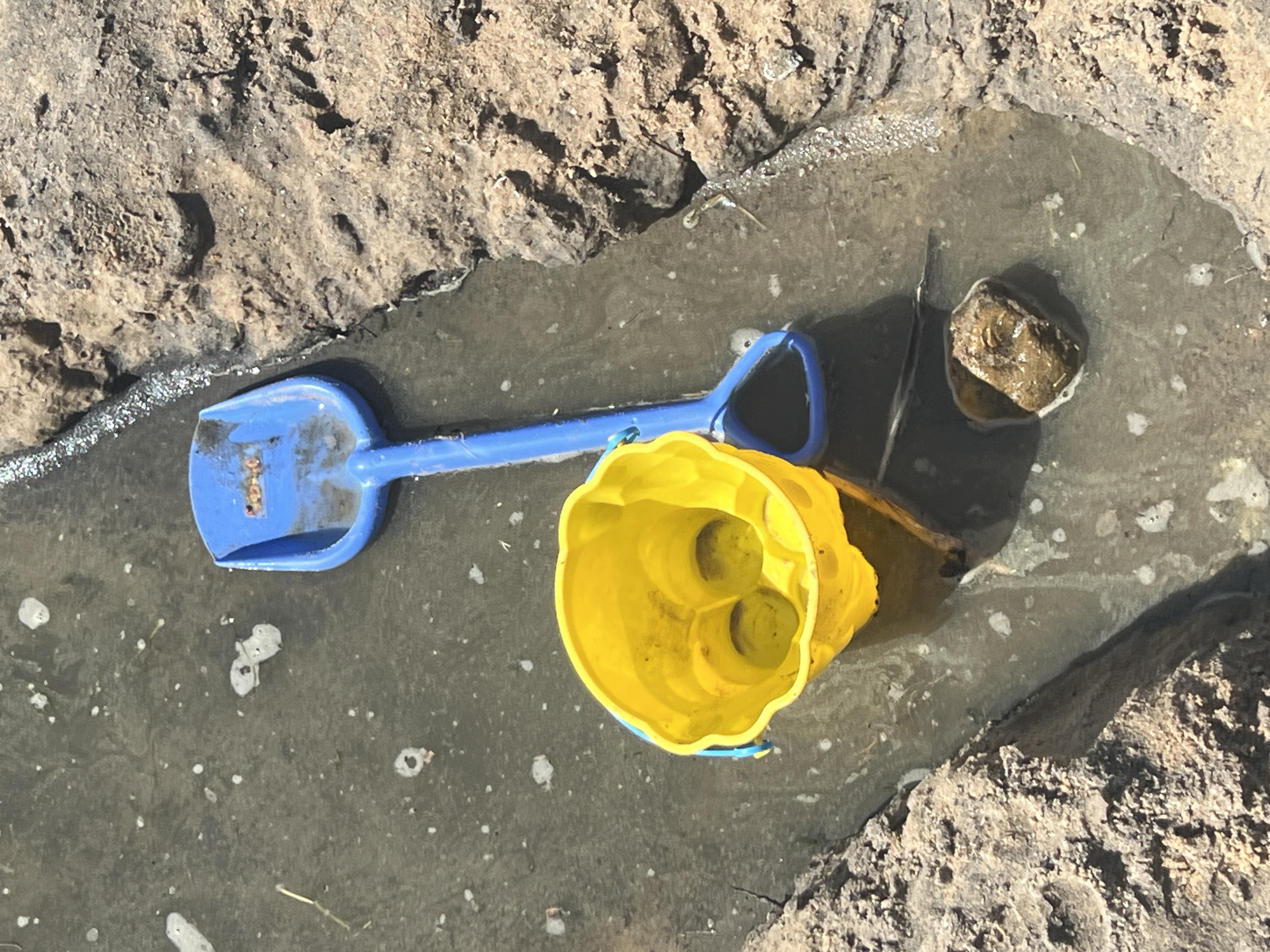 A blue plastic shovel and a yellow plastic bucket are submerged in shallow, murky water. The shovel is positioned diagonally with its handle pointing upwards and to the left. The bucket is to the left of the shovel, upright and filled with water. The scene appears to be a muddy, outdoor environment, possibly a puddle or a shallow stream bed, with rough, textured ground on the right side. The water contains small white bubbles and debris. There is a small rock in the water near the bottom center of the image. The overall impression is that of a child's toys left behind in a natural, wet environment. The location is not identifiable as Minsk, Belarus.