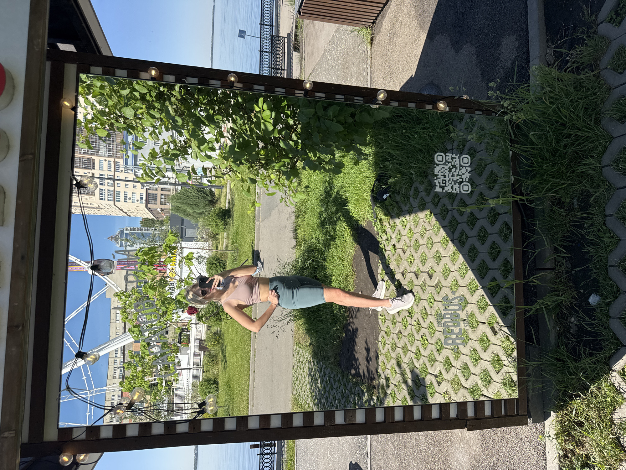 The image shows a woman in athletic attire taking a selfie in front of a mirrored structure. The structure is rectangular with exposed light bulbs along the top edge. The reflection shows a cityscape with a Ferris wheel and apartment buildings. The woman is wearing sunglasses, a light pink top, and greenish-blue shorts. The ground around the structure is paved with blocks and grass. A QR code is visible on the ground near the structure. The location is Saratov, Russia, evidenced by the Cyrillic lettering "РЕБУС" (Rebus) on the ground near the bottom edge of the mirror. A river with a decorative embankment is visible in the background.