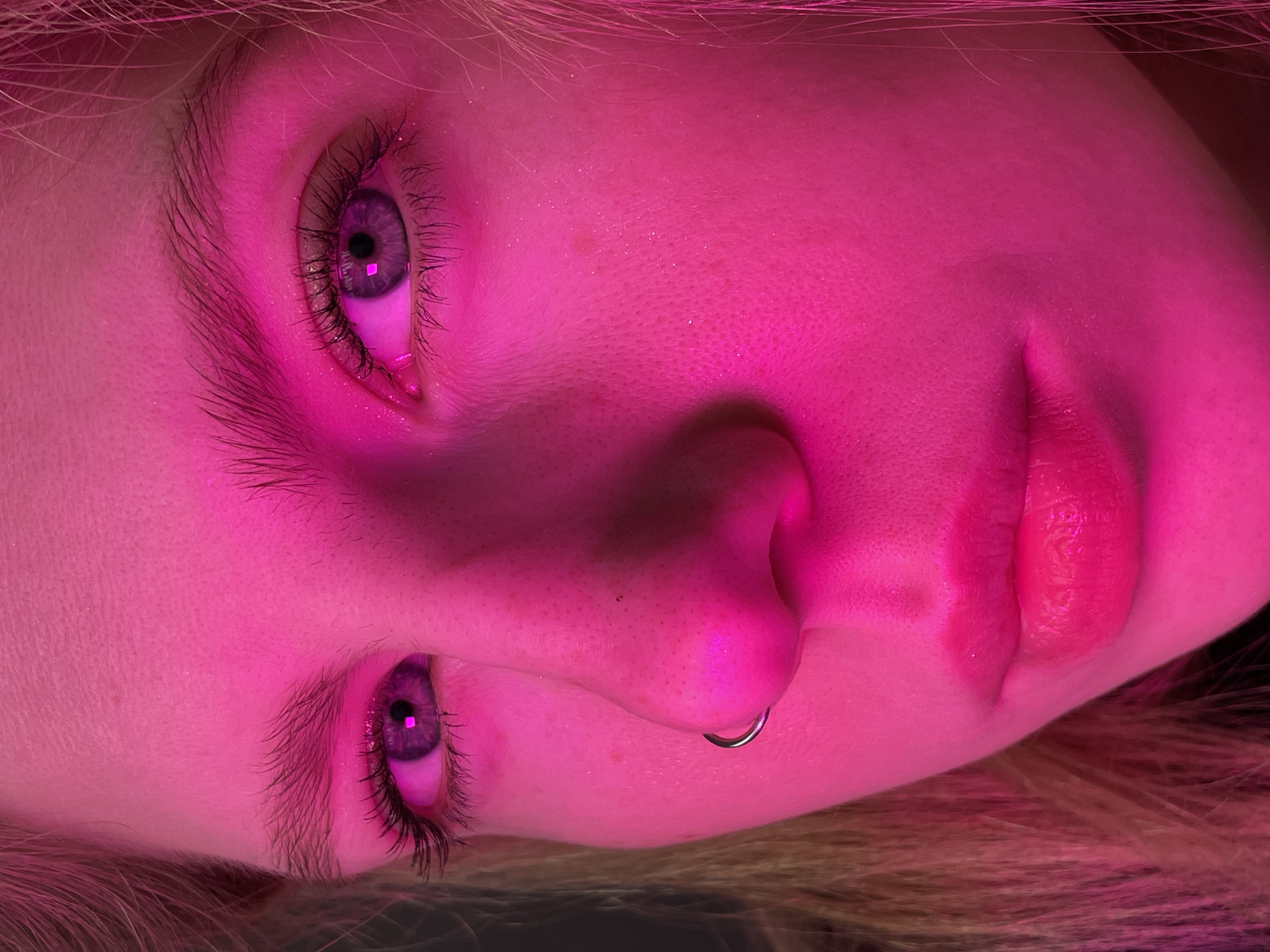 The media file displays a very close-up, horizontally oriented portrait of a young person's face, predominantly illuminated by a vibrant, intense pink light.
**Subjects:** The primary subject is a young person, depicted from the eyes down to the upper lip, with portions of their hair visible at the edges of the frame. Their eyes, which appear purple or lavender under the pink illumination, are open with visible eyelashes and catch a square reflection of the light source. The nose is prominent, featuring a silver-colored septum ring. The lips are full, slightly parted, and also reflect the strong pink light. The skin's texture, including pores and fine hairs, is detailed due to the close proximity.
**Objects:** The most notable object is the silver-colored, circular septum piercing worn in the subject's nose.
**Activity or event taking place:** The person appears to be posing for a photograph. No other specific activity is discernible; they are likely still or lying down, given the horizontal orientation of the face in the frame.
**Notable details:** The entire scene is drenched in a striking pink and magenta light, which colors the skin, hair, and even the natural eye color. The lighting creates a high-contrast, atmospheric, and possibly surreal aesthetic. There is no indication of time of day or weather. No location cues are visible within the image itself. The subject's expression is neutral, possibly contemplative or serene. There is no visible text.