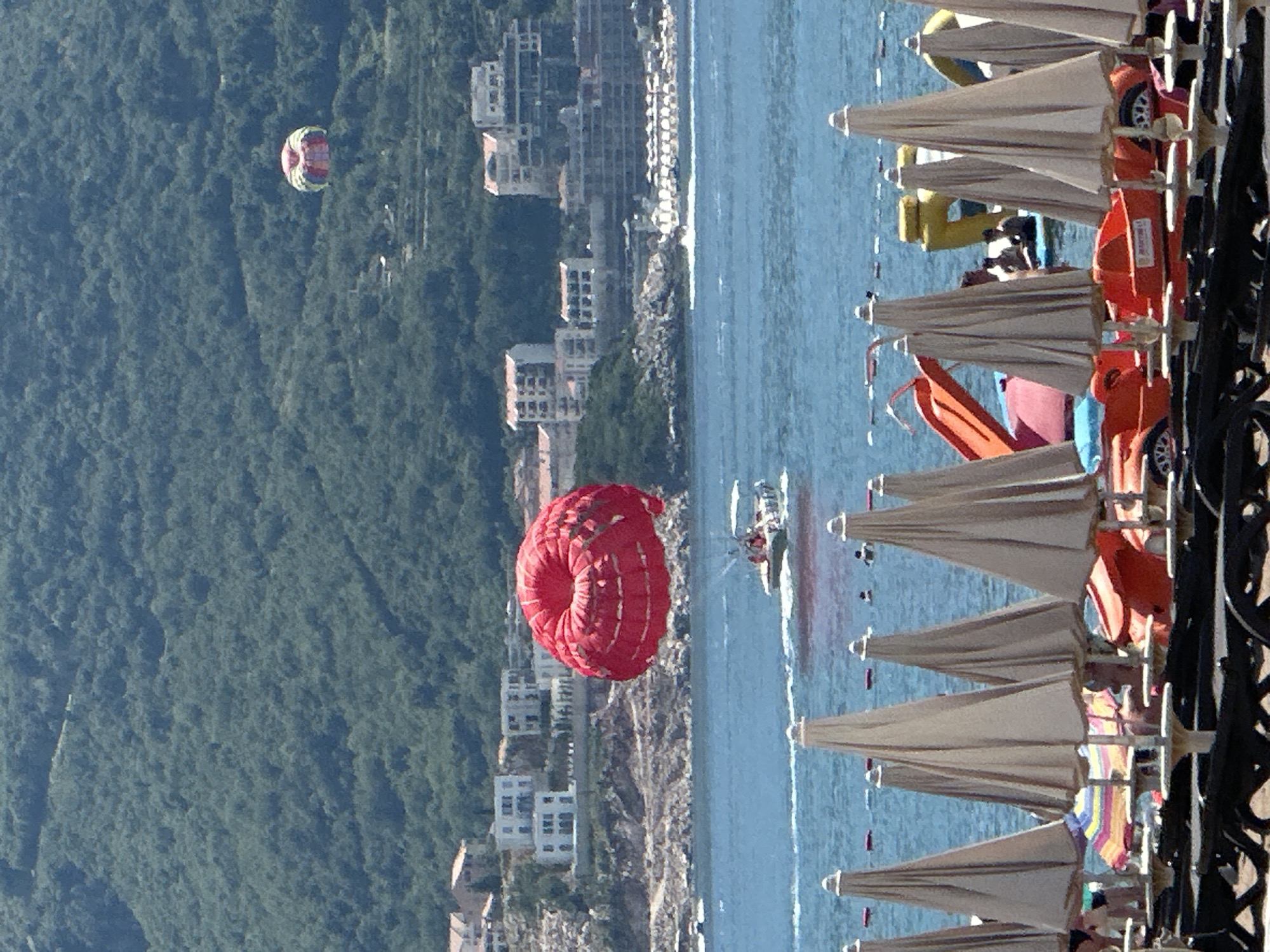 In Boreti, Montenegro, on a sunny day, a paraglider attached to a boat is seen soaring over the Adriatic Sea. The red parachute is prominent in the foreground, while the boat creating it is visible below. Further in the background, a smaller, multi-colored parachute is also visible in the air. The coastline is lined with buildings and lush green hills, indicative of a coastal resort town. The foreground of the image shows a beach with rows of closed beach umbrellas and lounge chairs, suggesting it is early or late in the day or that these are preparations for visitors. Some beach toys, including a red slide and pedal boats, are in the shallow water.