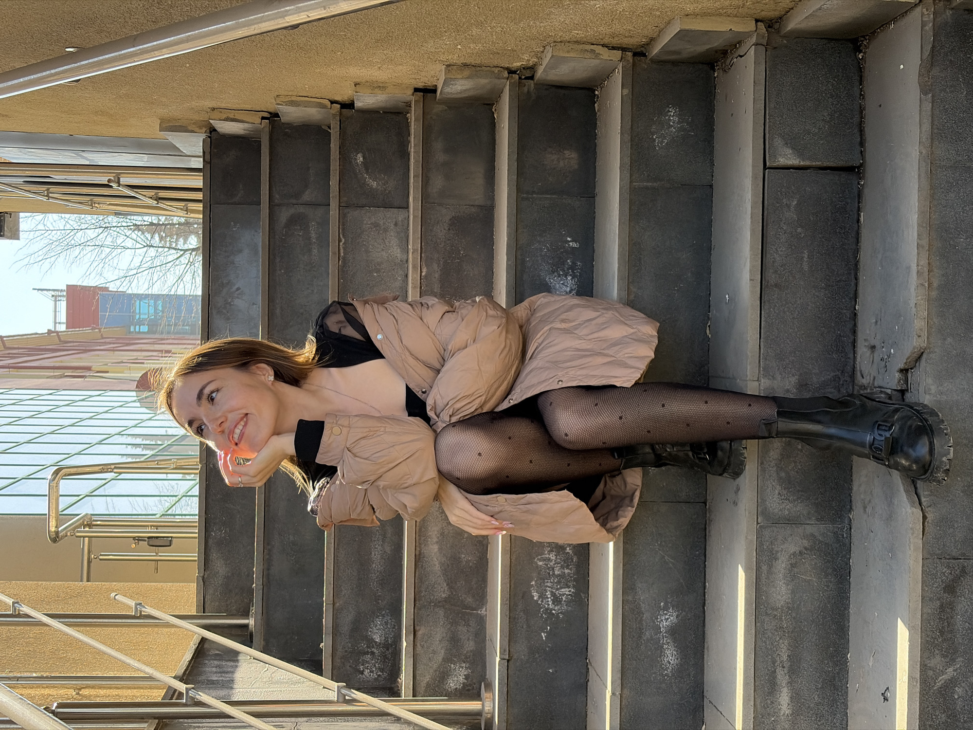 A young woman is lying on a set of concrete stairs with metal railings. She is wearing a tan puffer jacket, black fishnet stockings, and black boots. She is smiling and looking to the left. In the background is a building with glass windows and a red and blue structure. This image was taken in Izhevsk, Russia.