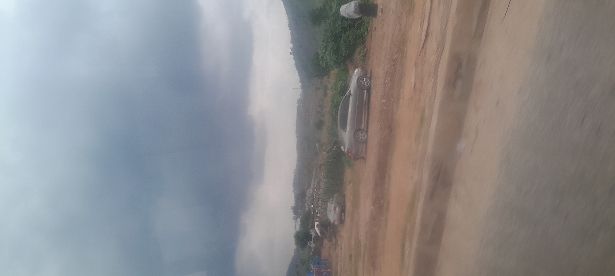 The image shows a semi-arid landscape with a grey, overcast sky. In the foreground, a dirt road runs diagonally across the frame. Two cars are visible on the dirt road. The car on the left is a light-colored sedan. The car on the right is a darker-colored sedan with a person partially visible leaning against it. In the background, there are some structures that appear to be makeshift dwellings or market stalls, with some vegetation and rolling hills. A large, dark cloud formation dominates the upper portion of the image, suggesting impending rain or stormy weather.
