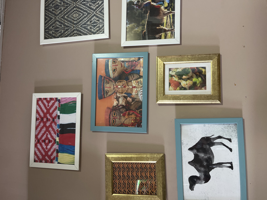 The image shows a wall with six framed pictures. The wall is painted a light brown color. The top left picture is a white frame with a picture of colorful woven fabric. The bottom left picture is a blue frame with a black and white silhouette of a camel. The top right picture is a white frame with a black and white patterned image. The picture directly below this frame is a white frame containing a picture of a llama. The picture below the top left picture is a light blue frame with an orange backdrop and three doll-like figures with colorful clothing. The last picture on the bottom right is a gold frame with a picture of a food dish. This appears to be a wall in a home or business located in Las Palmas de Gran Canaria, Spain at coordinates: latitude: 28.146, longitude: -15.43. The pictures are hung with visible white nail holes, though the placement is otherwise orderly and aesthetically pleasing. The pictures appear to be of diverse origin and theme, including traditional clothing, animals, food, and folk art.
