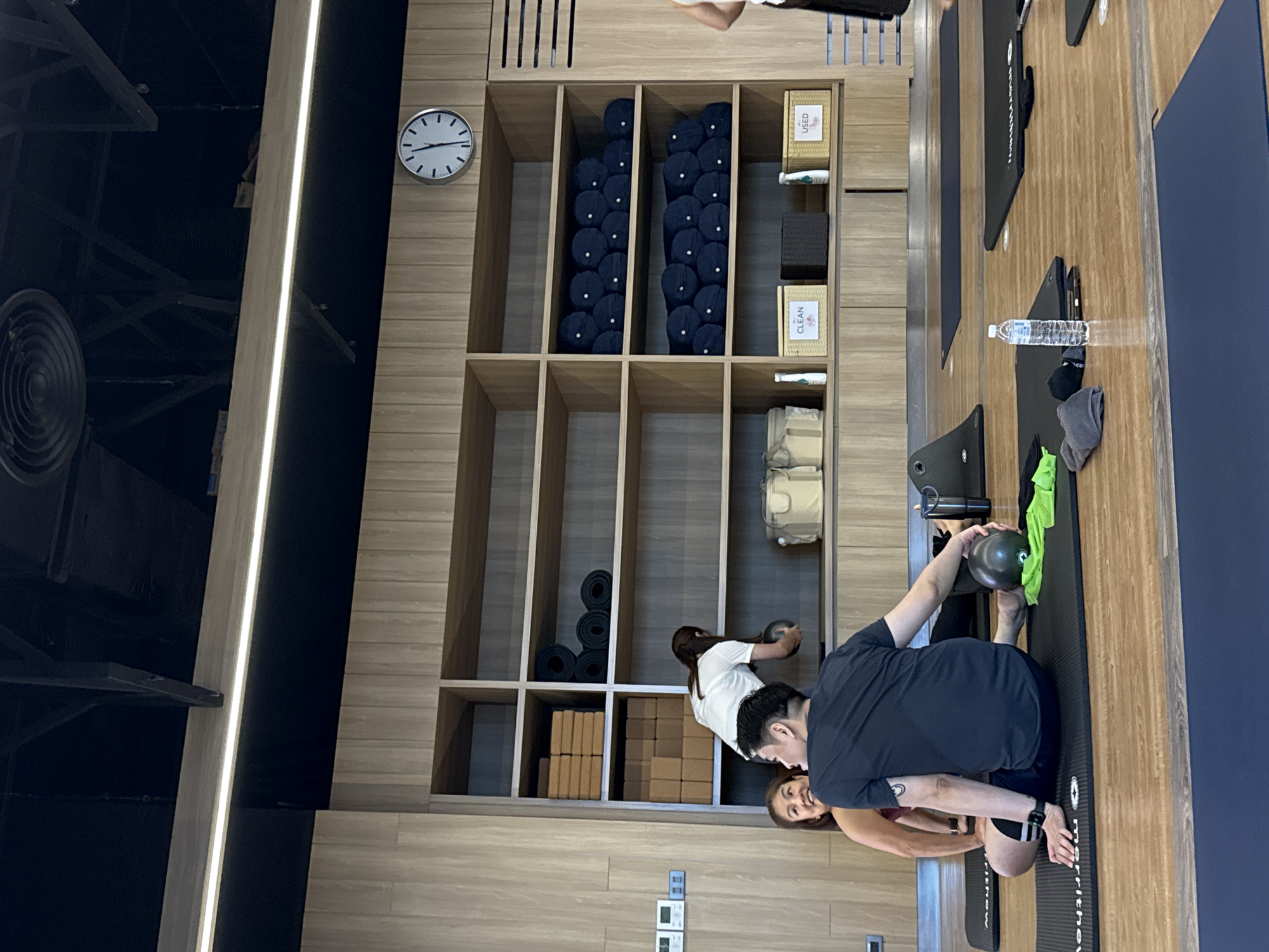 The image depicts an indoor yoga studio with wooden paneling and shelves. In the foreground, a man in a black t-shirt and shorts is seated on a yoga mat, holding a small black ball. A woman in a maroon top is to his left, also on a mat, smiling. Behind them, a woman in a white t-shirt is reaching for a dark ball on a shelf. The shelves contain yoga blocks, rolled-up yoga mats, and storage bins labeled "CLEAN" and "USED". A clock is visible on the wall above the shelves. The floor is wooden, and several yoga mats are laid out. The overall environment suggests a class or practice session.
