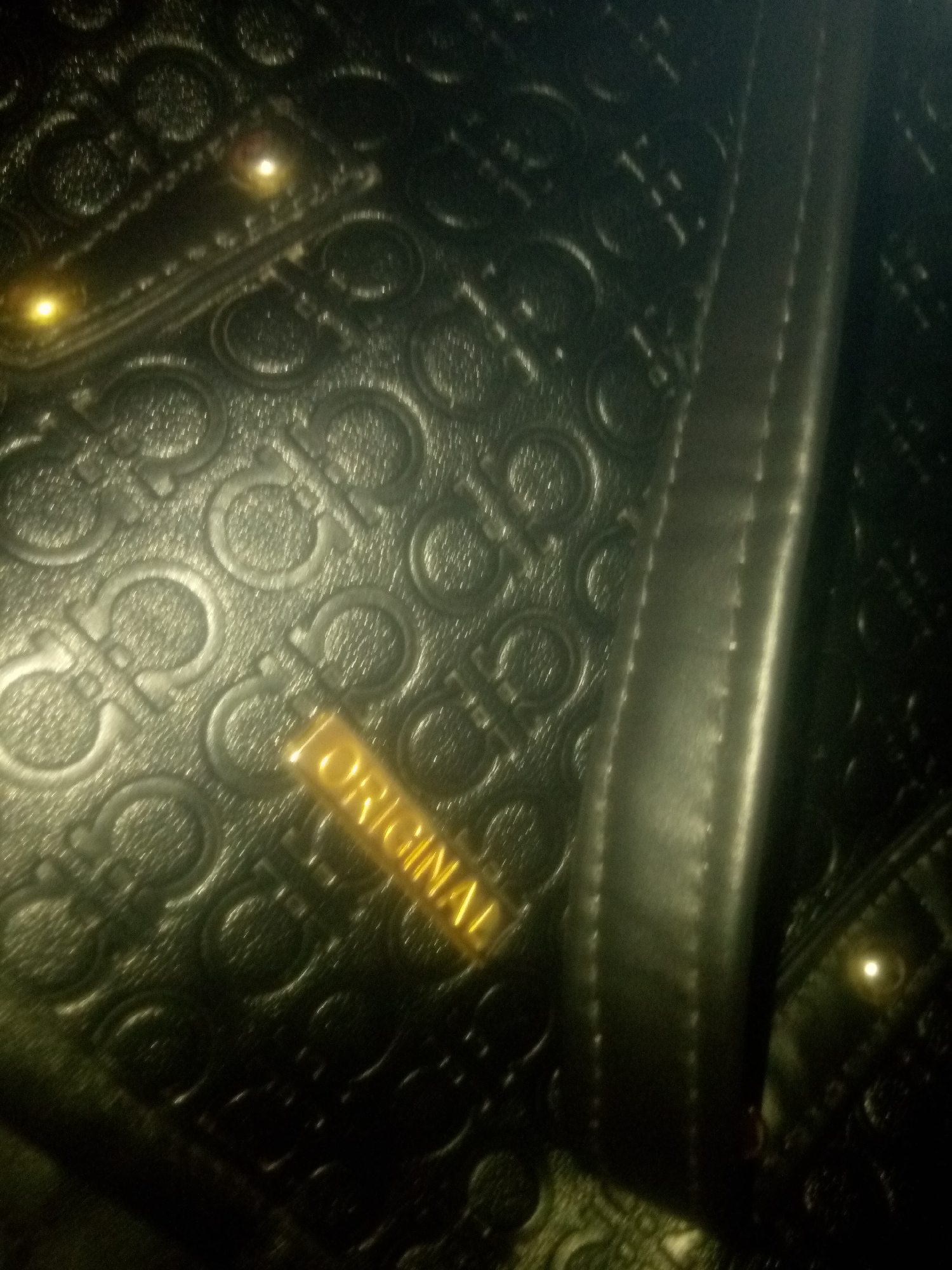 The image shows a close-up of a black textured material, likely a handbag or wallet. The material has an embossed pattern of interlocking "C" shapes. A gold-colored rectangular tag with the word "ORIGINAL" embossed on it is diagonally placed across the lower half of the image. The lighting is dim and creates reflections and highlights on the glossy surfaces of the material and the tag. The background is dark and out of focus. There are no people or signs of a specific city location.