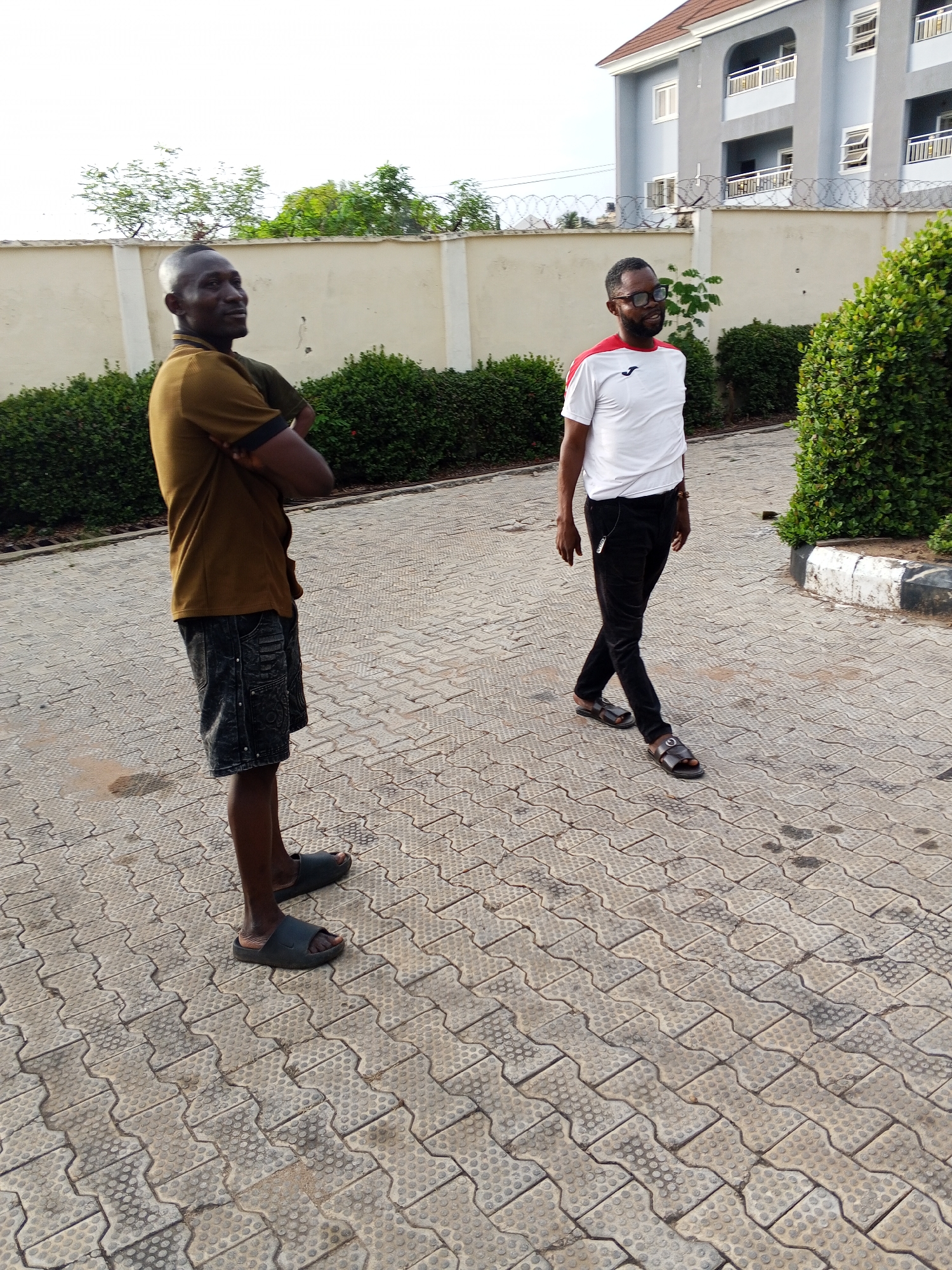 Two men are standing on a paved area. The man on the left is wearing a brown shirt, patterned shorts, and sandals. He has his arms crossed. The man on the right is wearing a white and red shirt, black pants, and sandals. He is walking towards the right. A low wall is behind the men with green bushes planted along it. A building is visible in the background with barbed wire on top of the wall. The scene appears to be a driveway or parking area. The location could be Lokoja, Nigeria.