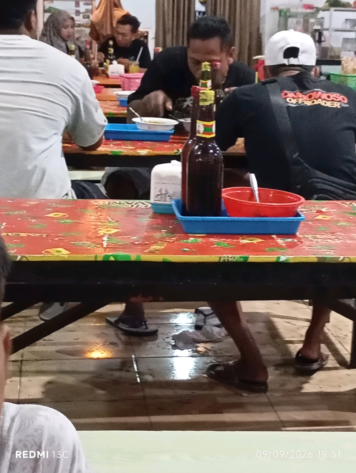 The image depicts a dimly lit interior of a restaurant in Wuluhan, Indonesia, likely a local eatery or warung. Several individuals are seated at tables, suggesting a casual dining environment. On one table, a red and yellow patterned tablecloth is visible, adorned with a blue tray holding a red bowl with a spoon and a dark glass bottle. Another table has a similar tablecloth, with a white container and a blue plastic tray on it. People are dressed casually, with some wearing t-shirts and one individual in the foreground wearing a white shirt. The flooring appears to be tiled and wet in some areas, possibly indicating a recent spill or cleaning. The overall ambiance suggests a relaxed, communal gathering or meal.