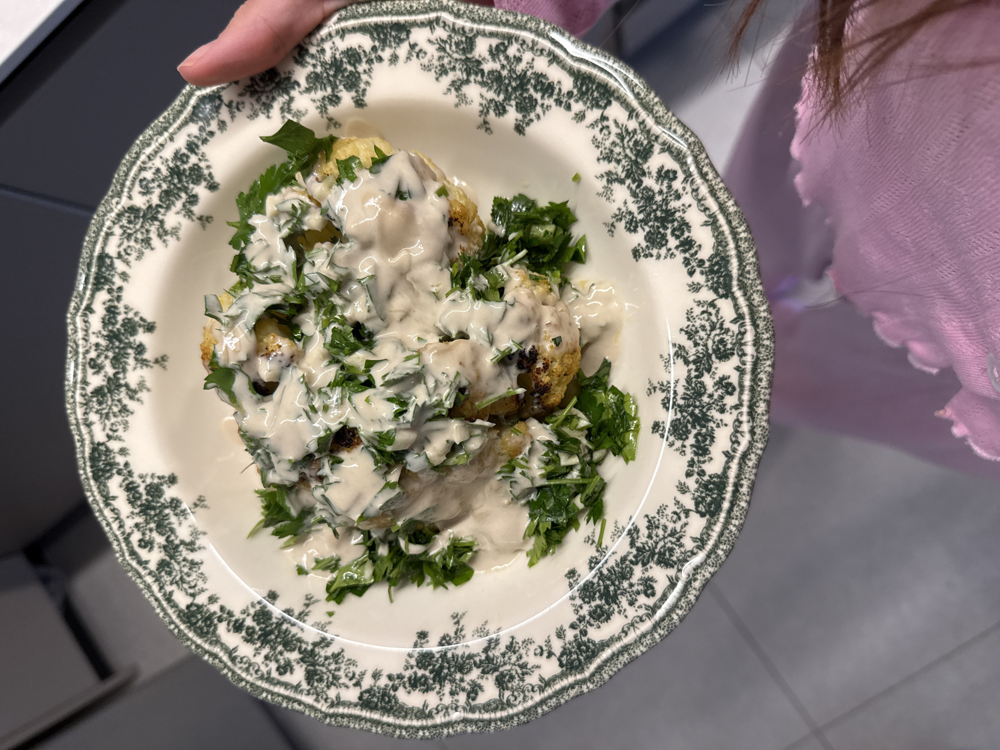 A person holds a bowl of food. The bowl is white with a green floral pattern along the rim. Inside the bowl are several pieces of food, potentially fried or roasted, covered in a white sauce and garnished with chopped green herbs, likely parsley. The person holding the bowl is wearing a pink sweater or garment. The background is blurred, but hints of a kitchen setting are visible. The location is unspecified but indicated to be Givatayim, Israel.