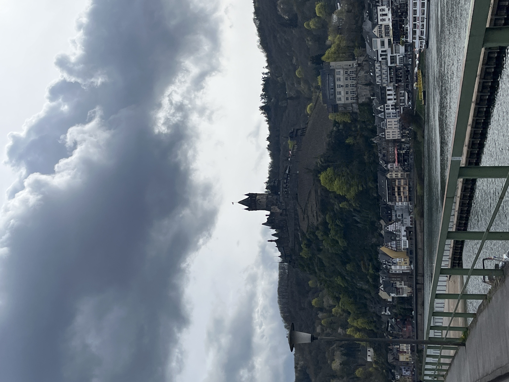 The image presents a landscape in Cochem, Germany, showcasing Reichsburg Cochem, a medieval castle atop a vine-covered hill overlooking the town. The Moselle River flows alongside a densely populated urban area, characterized by various buildings and structures. A bridge with green metal railings is situated in the foreground, partially obscuring the view. The sky is overcast with heavy, dark clouds. No people are visible.
