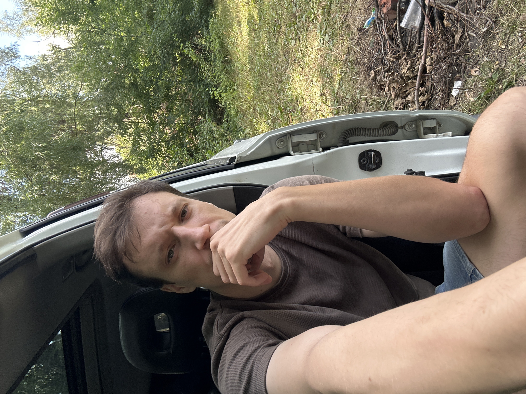 The image shows a man in his late 20s or early 30s sitting in the open driver's side of a white vehicle. He is wearing a dark brown short-sleeved t-shirt and denim shorts. His right arm is resting on his knee, and his left hand is raised to his chin, with his fingers touching his mouth. He has light brown hair and is looking directly at the camera with a pensive expression. The car appears to be parked outdoors in a natural setting, with trees and foliage visible in the background. The ground around the car is covered with dry leaves and twigs, suggesting a wooded or park-like area. There is a blue plastic object and a clear plastic cup visible on the ground to the right of the vehicle. The overall scene suggests a casual, outdoor setting. The location is identified as Makiv, Ukraine.