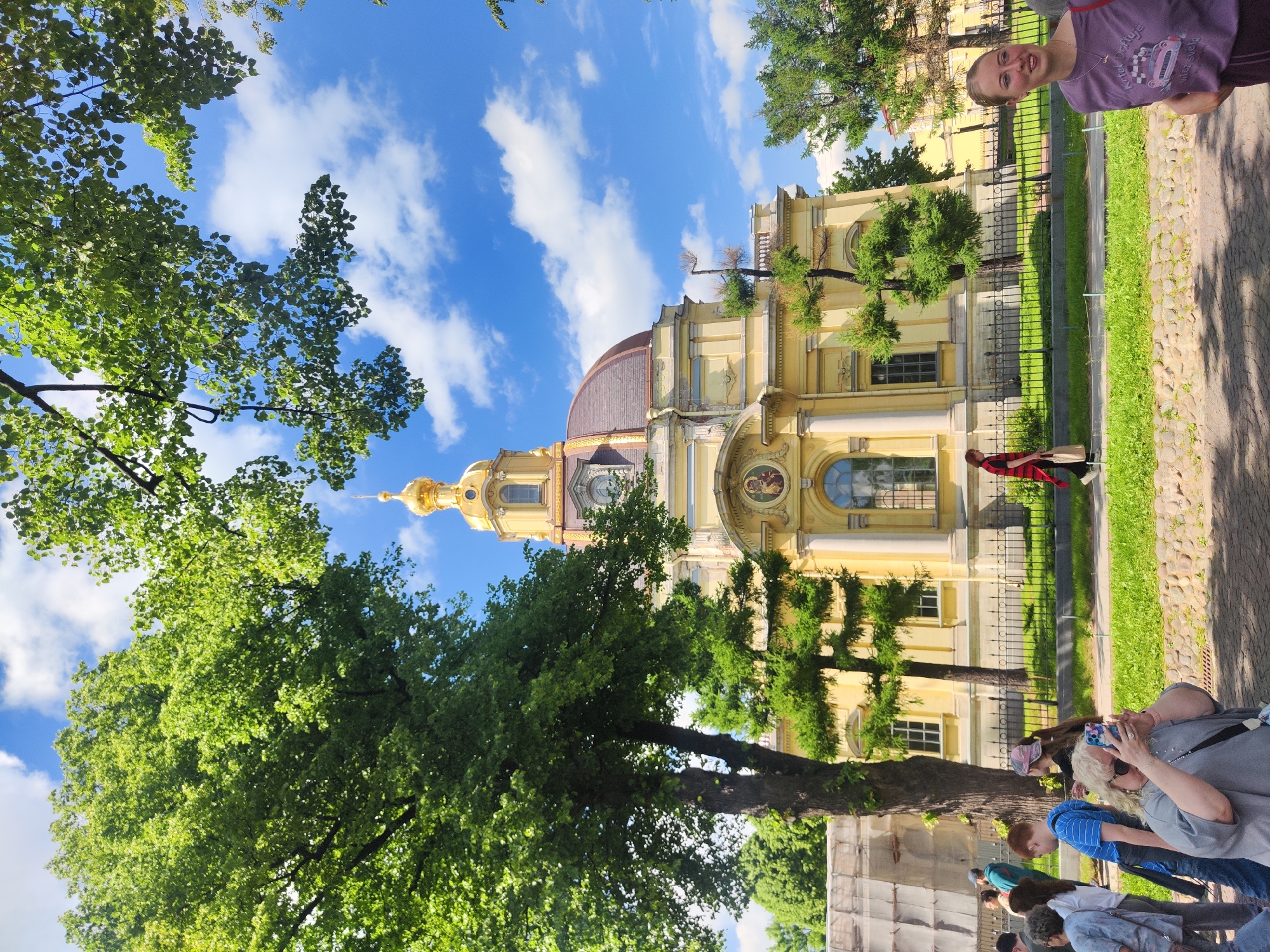 This image captures a sunny day with a bright blue sky and fluffy white clouds, framing a prominent yellow building with a golden dome and a reddish-brown dome, characteristic of Saint Petersburg's architectural style. The building appears to be a church or cathedral. In the foreground, several people are visible, some walking and others observing. One woman in a grey top is holding up a phone to take a picture, focused on the building. To the right, another young woman in a purple t-shirt with a graphic print is looking to the side, seemingly engaged in conversation or observing something outside the frame. Lush green trees with dense foliage are scattered throughout the scene, some providing shade and framing the top of the image. A black metal fence separates the paved walkway from a green grassy area in front of the building. The overall scene suggests a tourist attraction or a public space in Saint Petersburg on a pleasant day.
