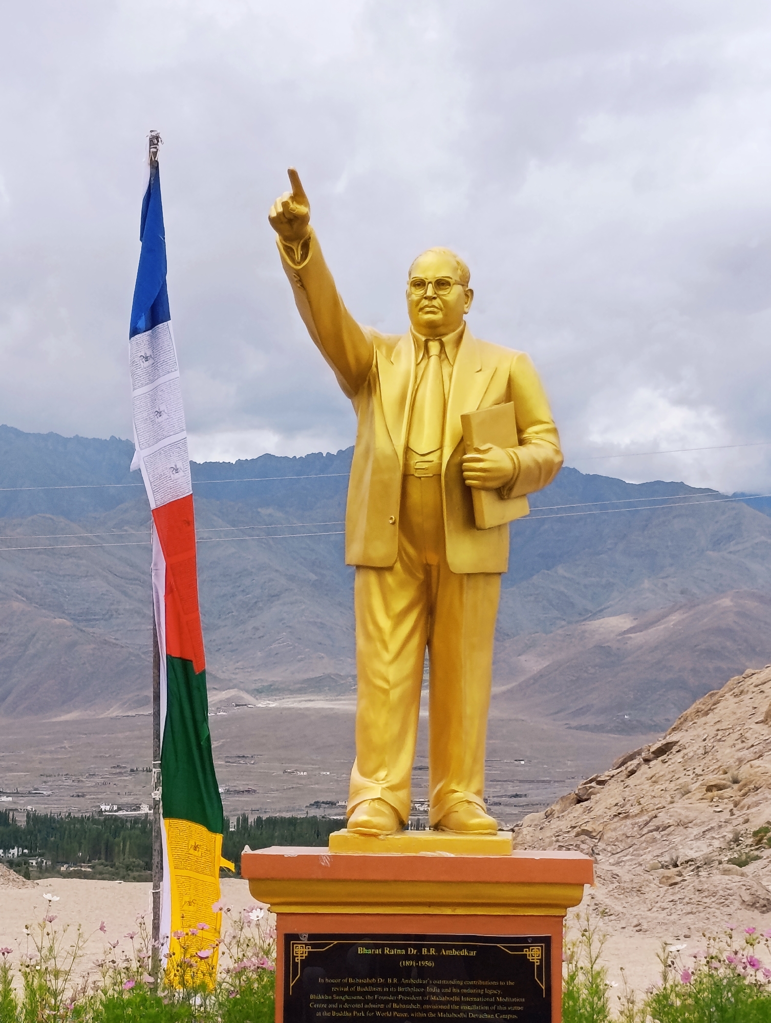 A full-body, golden statue of a man in a suit and tie stands on a pedestal in front of a mountainous landscape and a cloudy sky. The statue, which appears to be of Dr. B. R. Ambedkar, is located in Leh, India, at the statue of Buddha, Mahabodhi Meditation Centre, and the Mahabodhi Devasan Campus. To the left of the statue is a Tibetan prayer flag, displaying a variety of colors and Buddhist symbols. The ground in front of the pedestal is covered in dry grass and scattered pink flowers. In the background, a valley is visible, dotted with houses and trees. The mountains in the background are barren and rocky, with a dusty haze obscuring the far distance. The sky is overcast with dramatic, grey clouds.